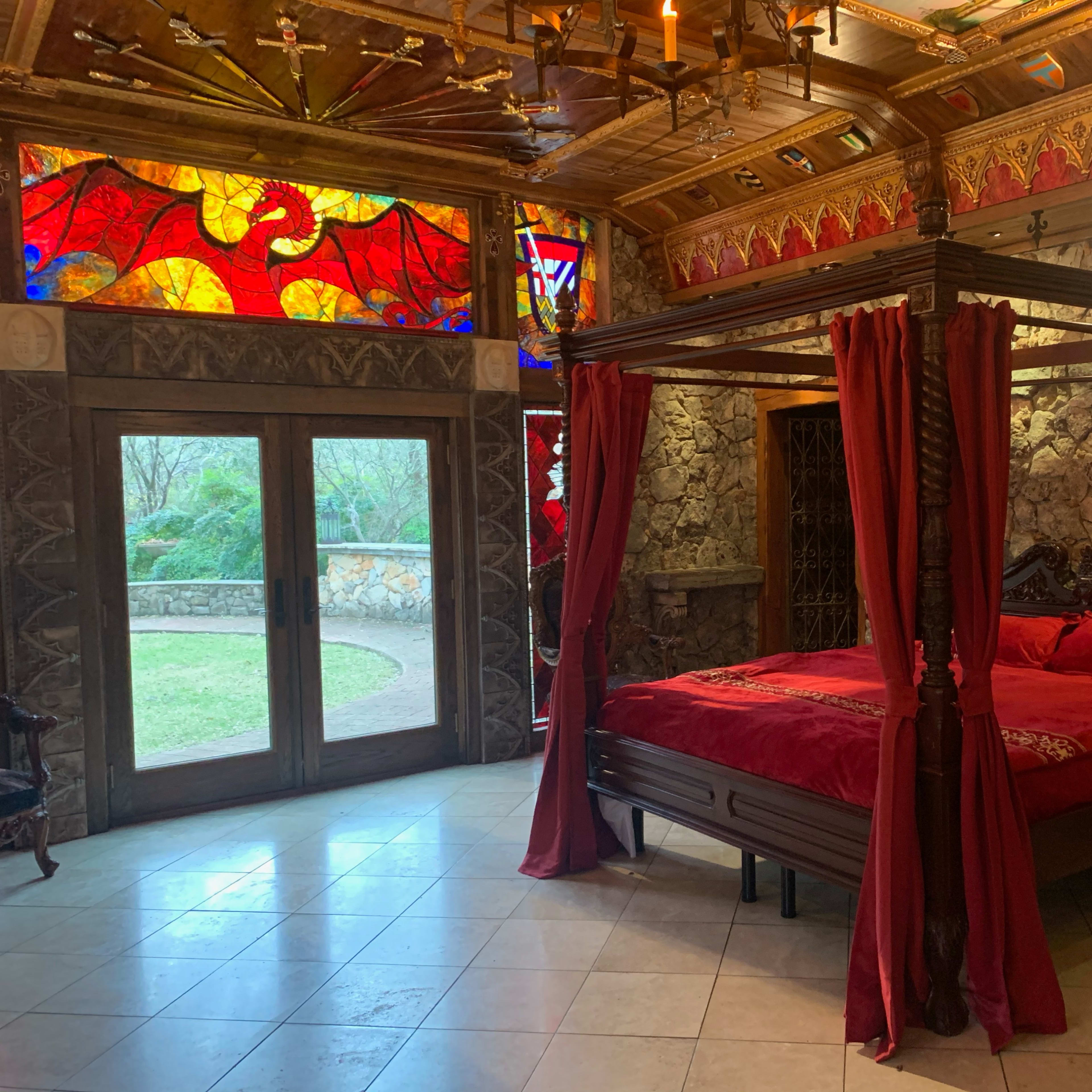 A four-poster bed with red drapes in a stone-walled bedroom featuring stained glass windows and a glass door leading to an outdoor area.