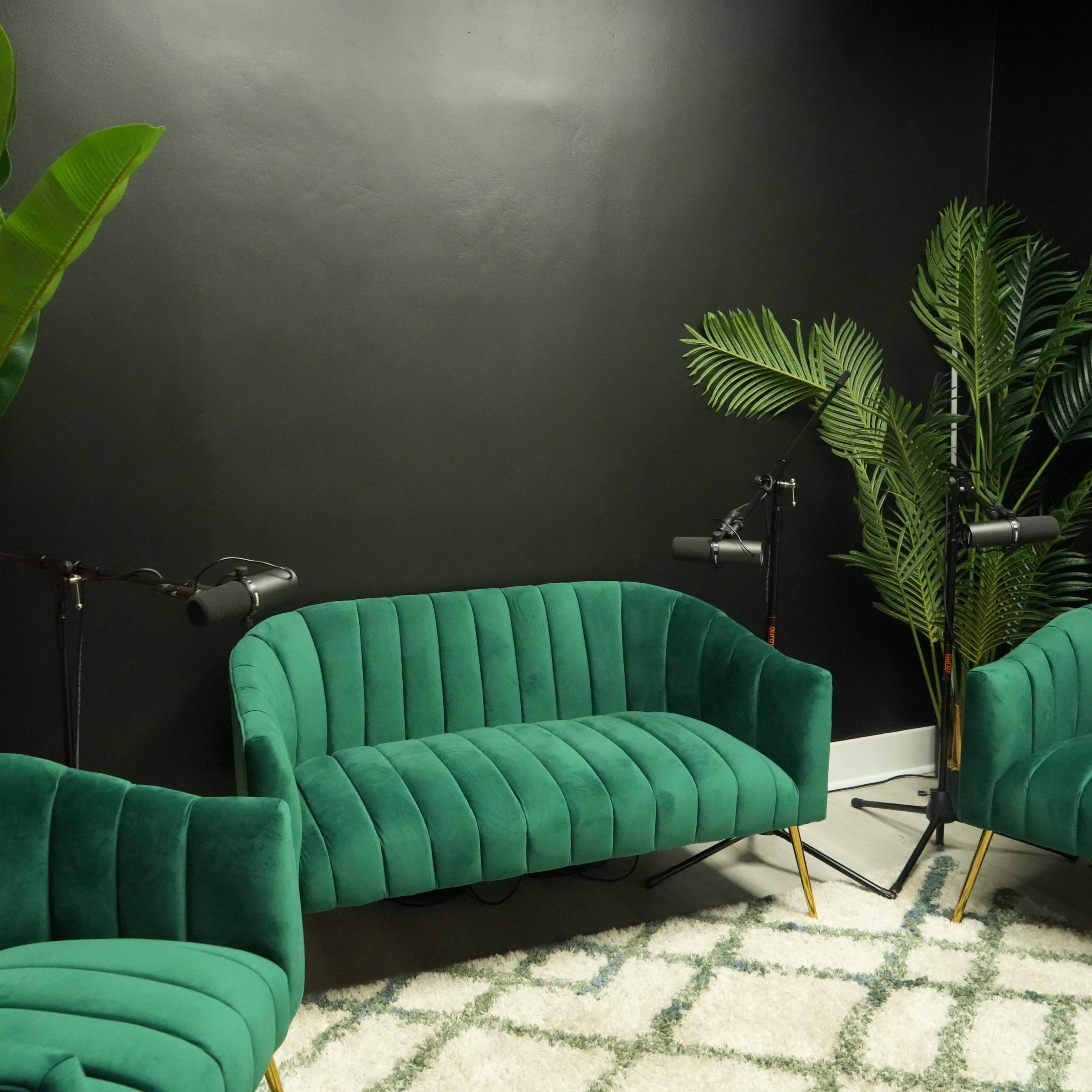 A cozy seating area with three green velvet chairs, a textured rug, and large potted plants against a dark wall.