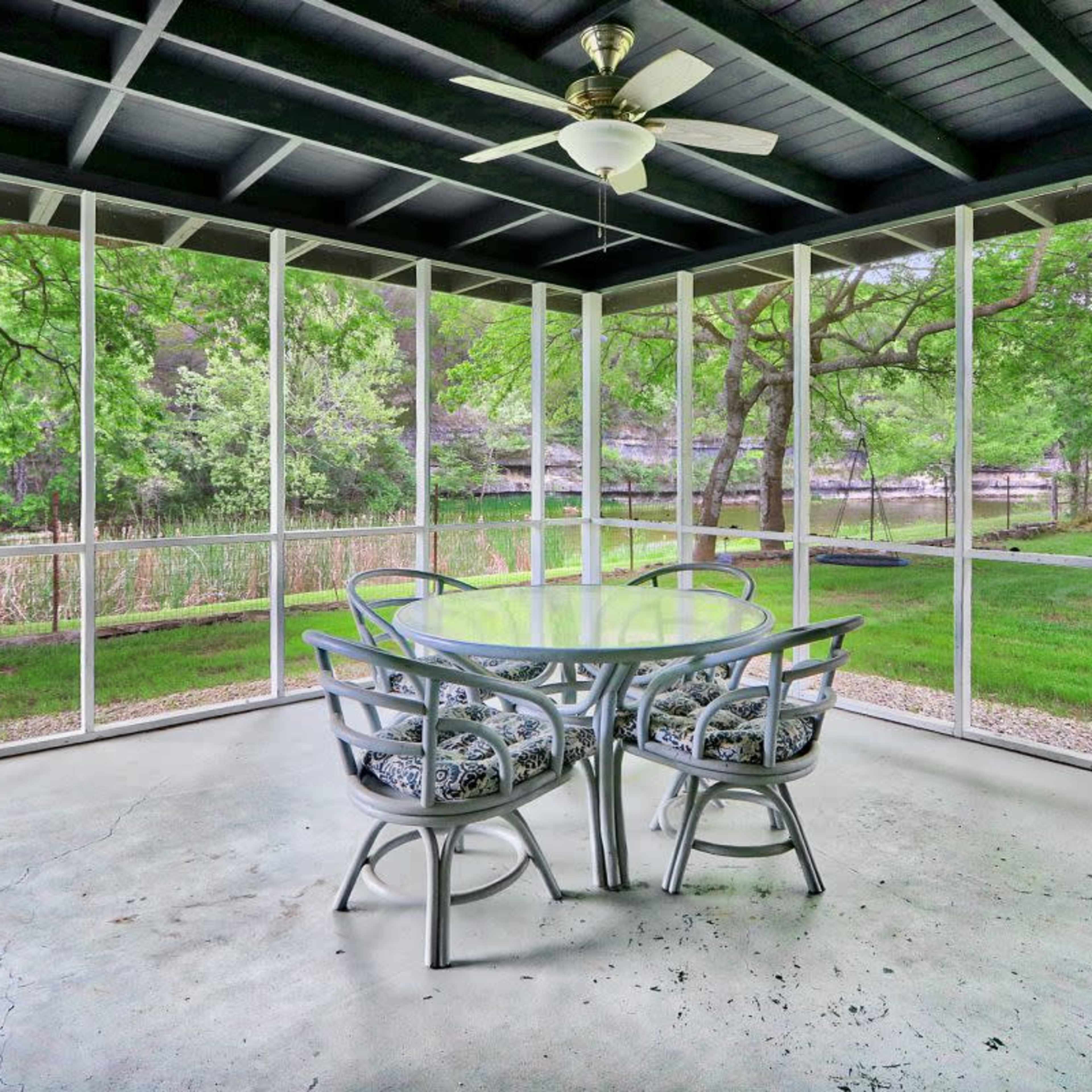 A screened-in porch featuring a glass table and four chairs overlooking a grassy yard with trees.