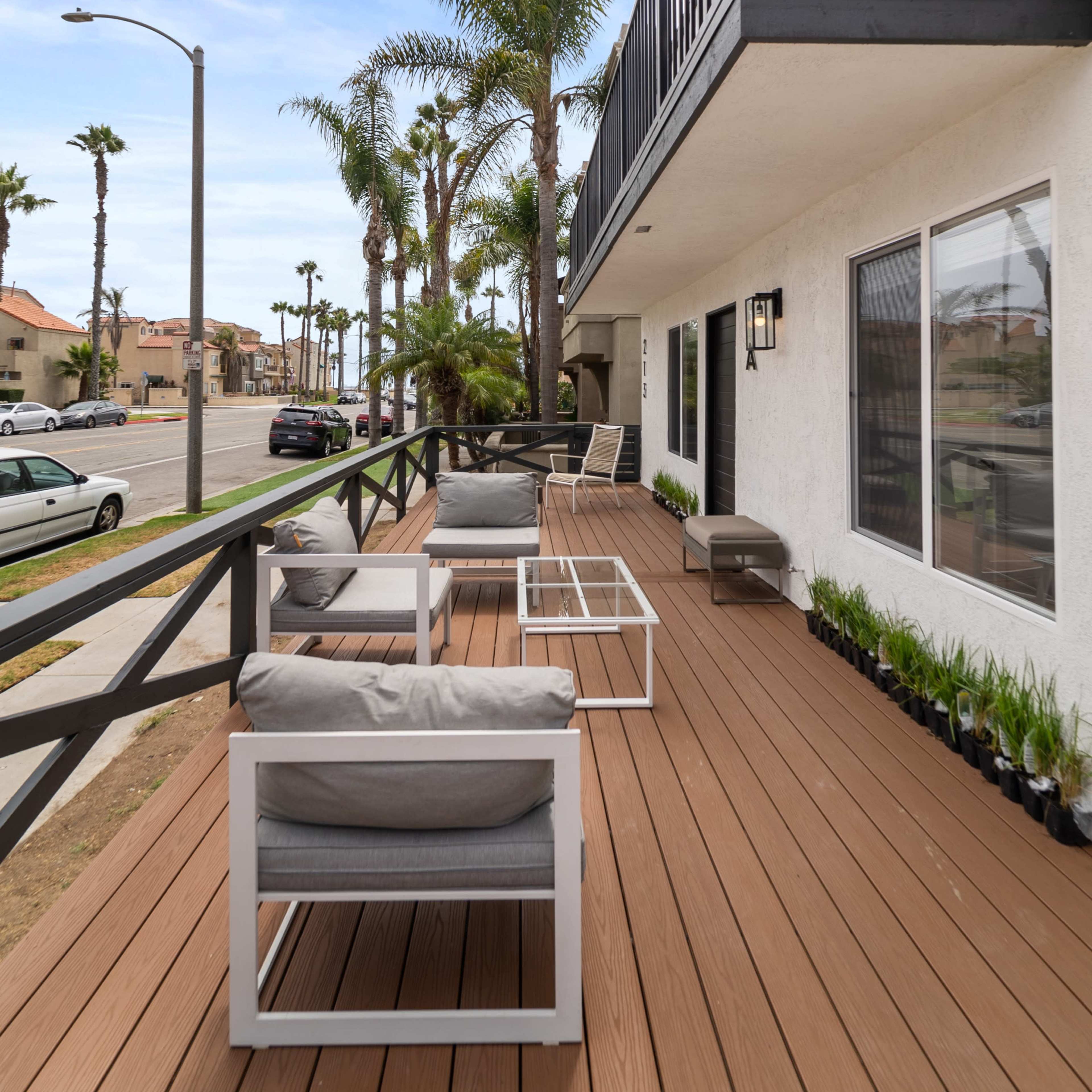 A wooden deck with modern furniture is located on the second floor of a building, surrounded by palm trees and facing a street.