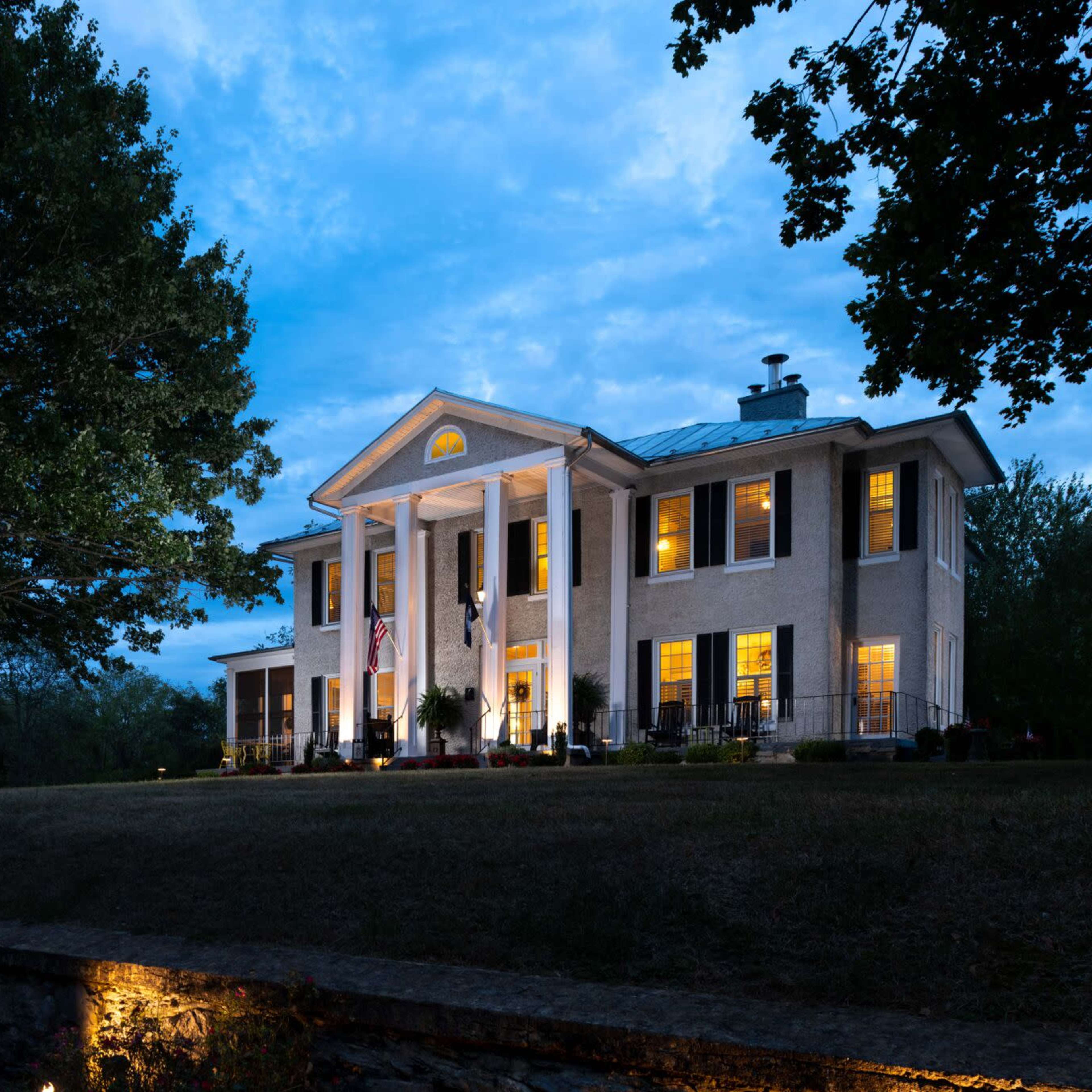 A large, two-story house with white columns is illuminated in the evening light, surrounded by trees and landscaped shrubs.