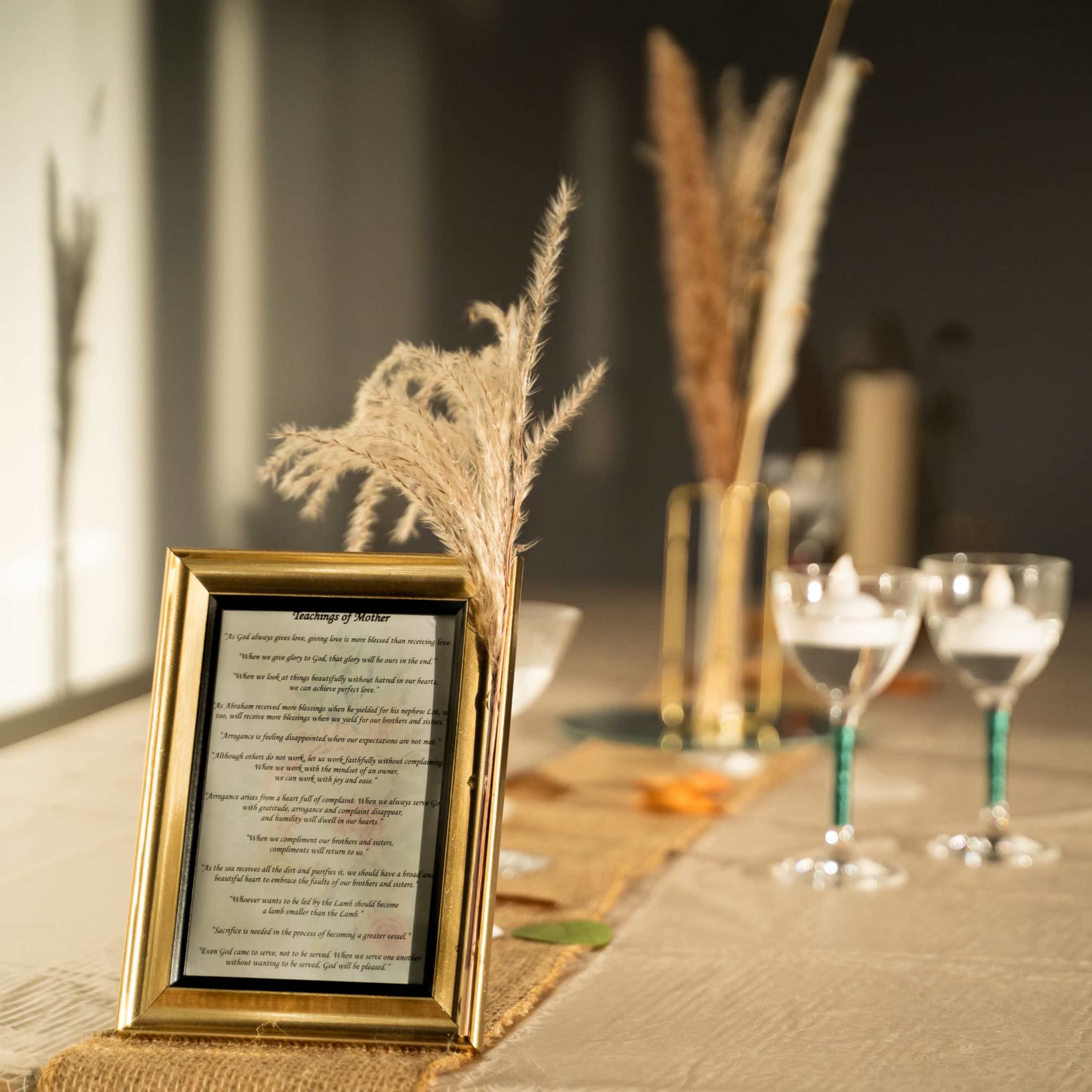 A framed document sits on a table adorned with decorative grasses and three elegant glasses filled with a liquid.