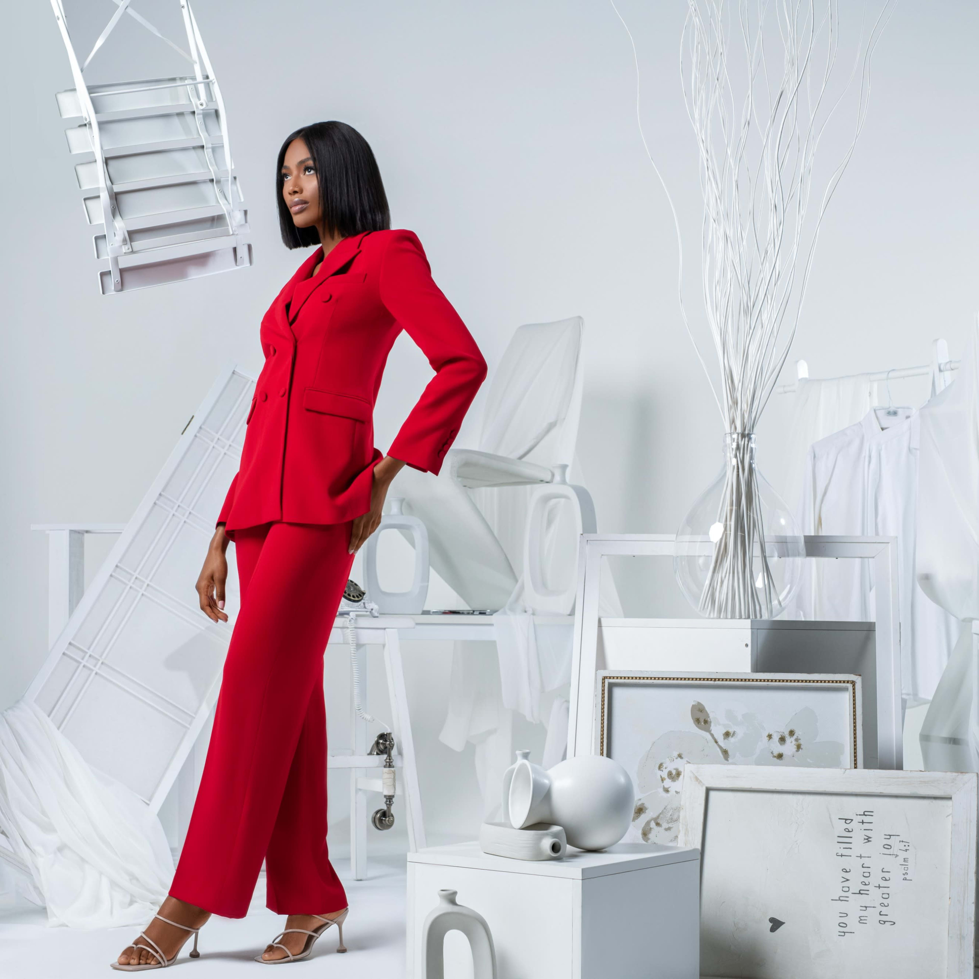 A model poses in a red suit among a minimalist white backdrop with various pieces of furniture and decor.