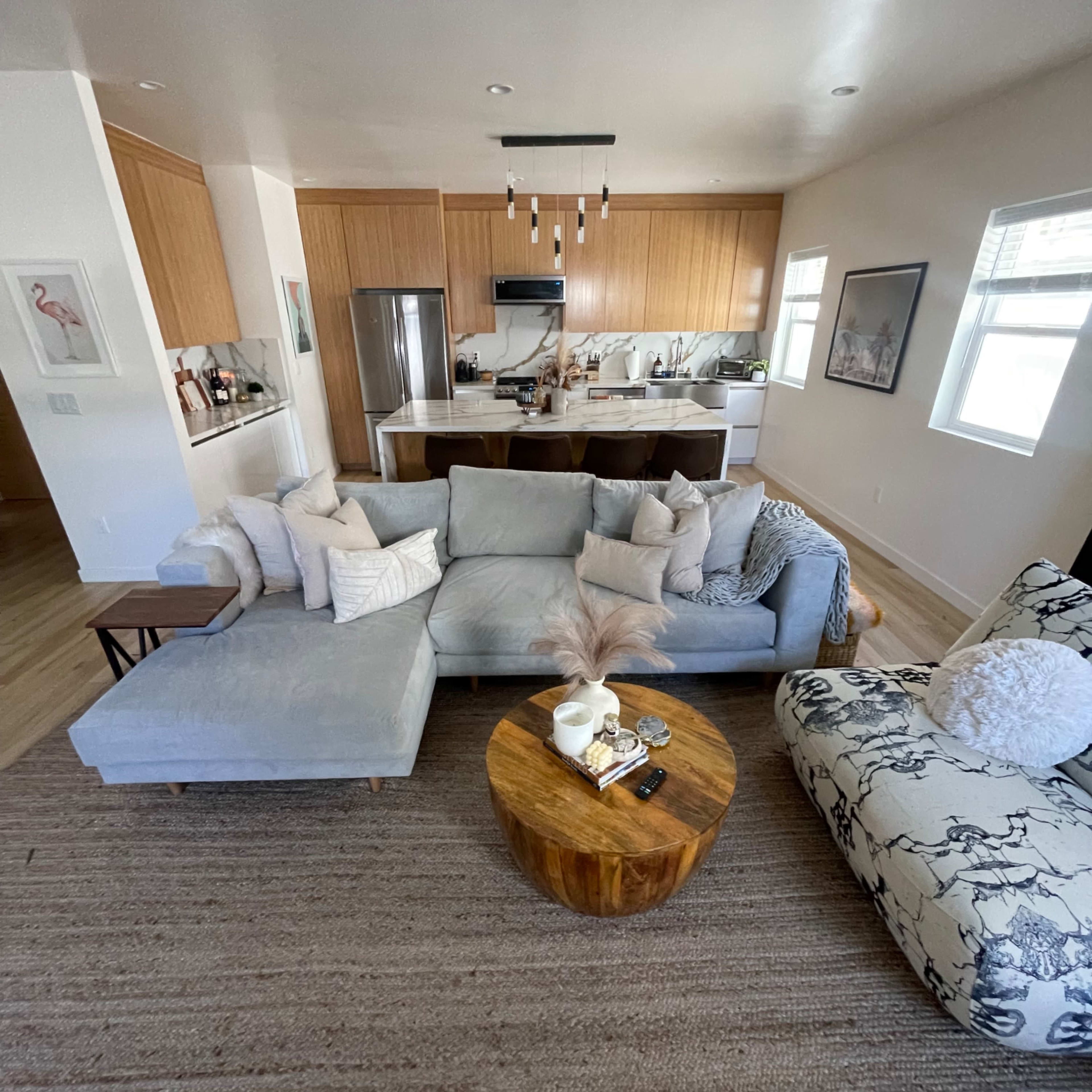 A living room features a sectional sofa, a round wooden coffee table, and a patterned armchair, with a view of an open kitchen in the background.