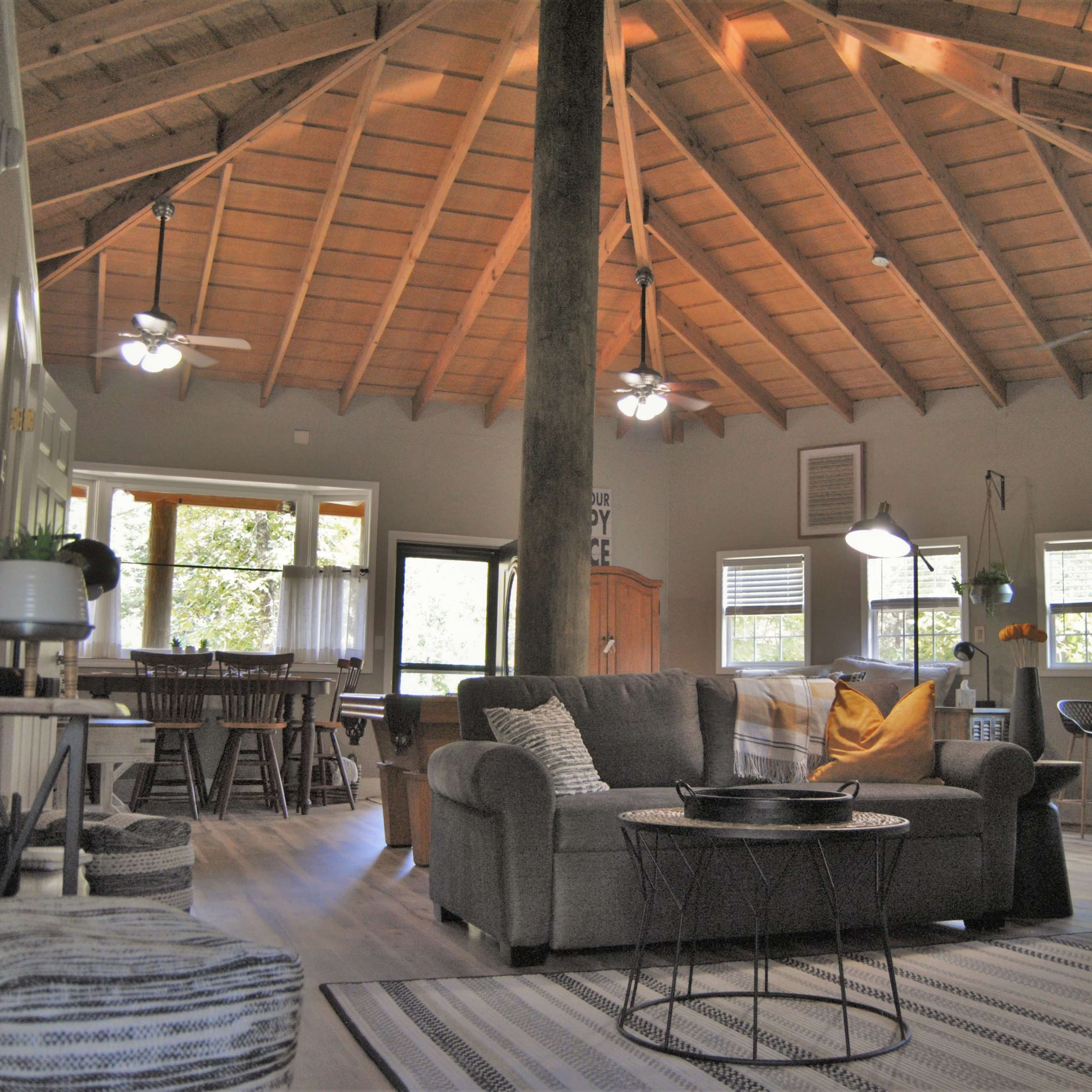 An open-concept living space with a high, wooden vaulted ceiling, featuring a gray sofa, a coffee table, and a dining area with a large window.