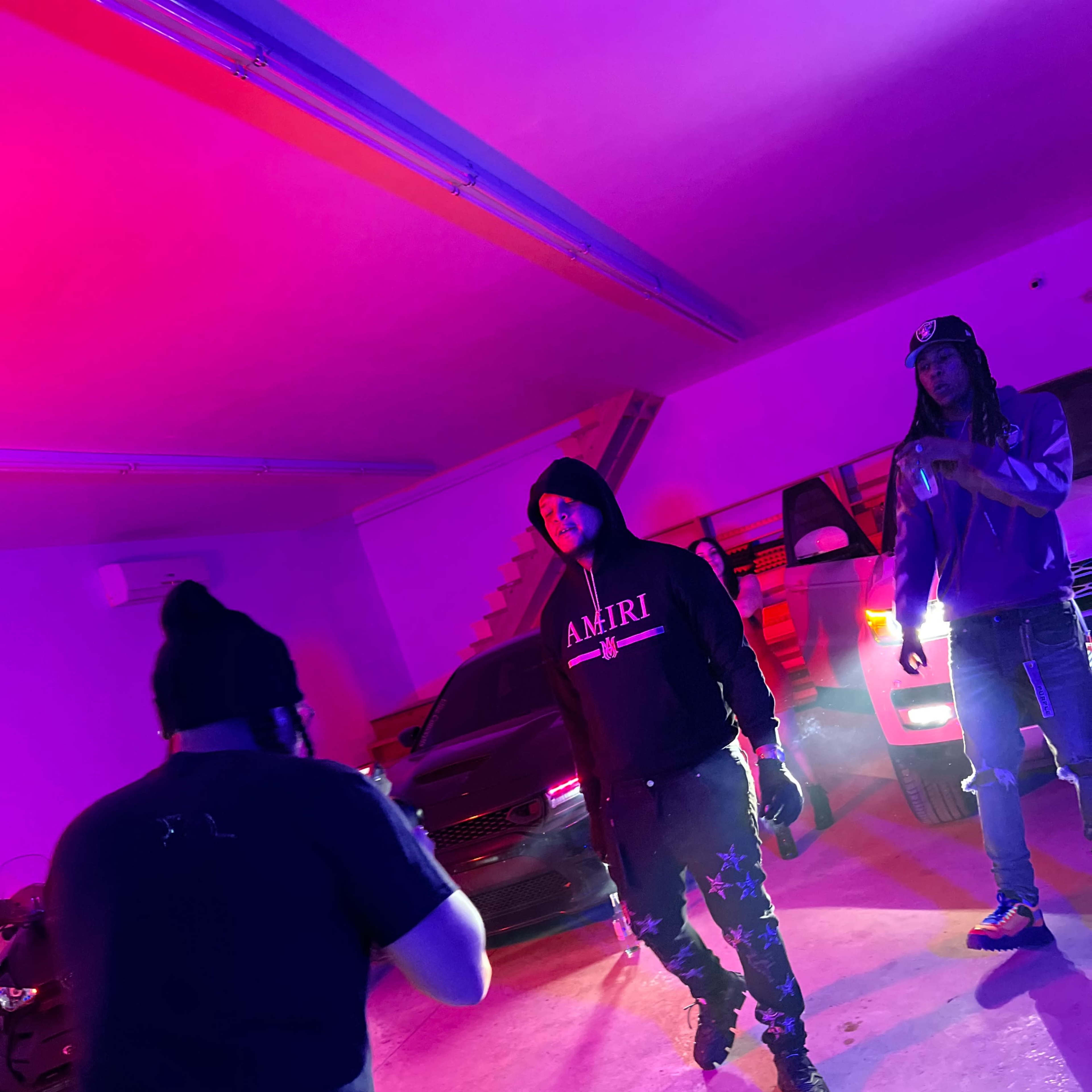 Two individuals in hooded sweatshirts standing in a dimly lit garage with colored lighting, while a third person appears to be taking a photo or video of them.