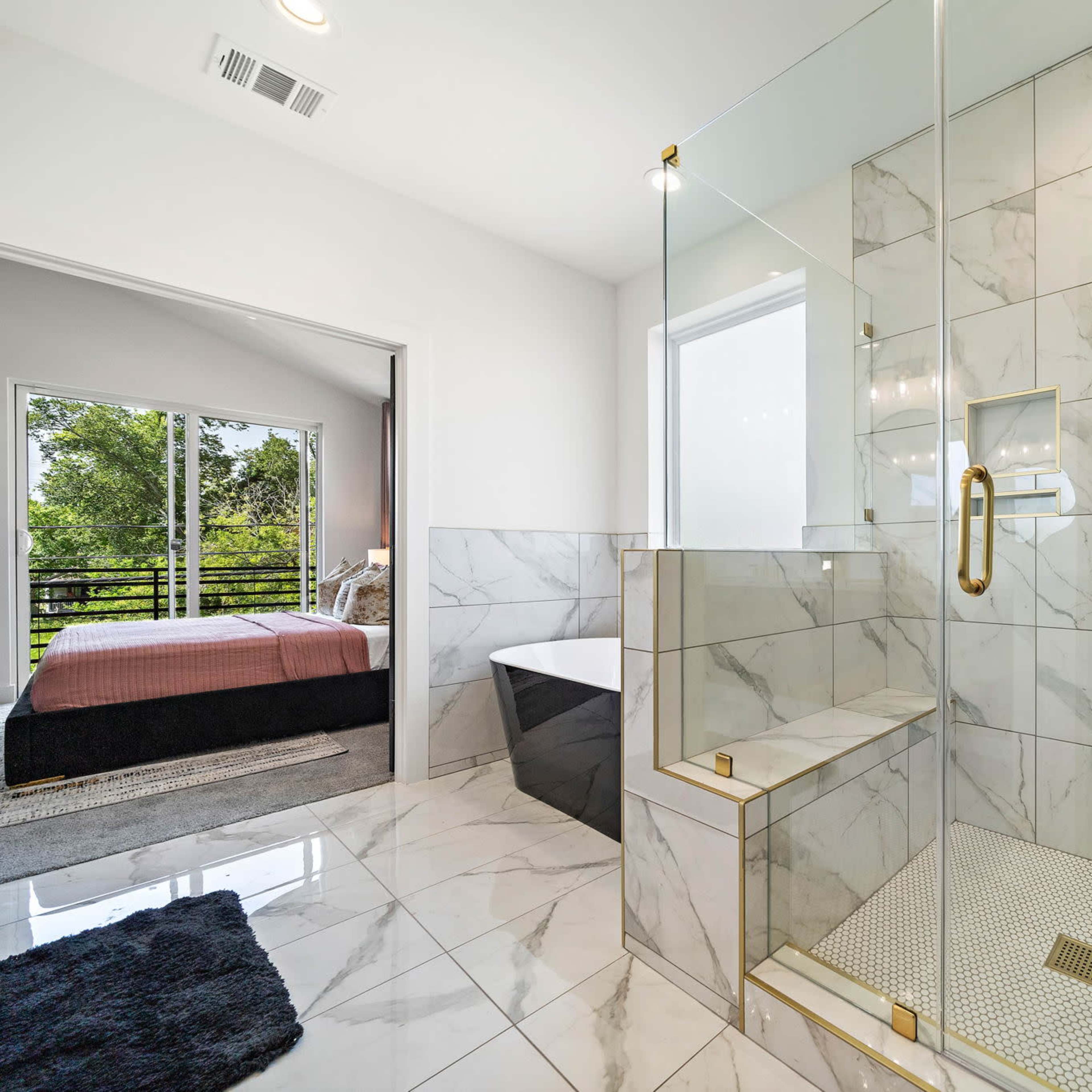 A modern bathroom featuring a glass shower, marble tiles, a freestanding bathtub, and views of an outdoor area through a large window.