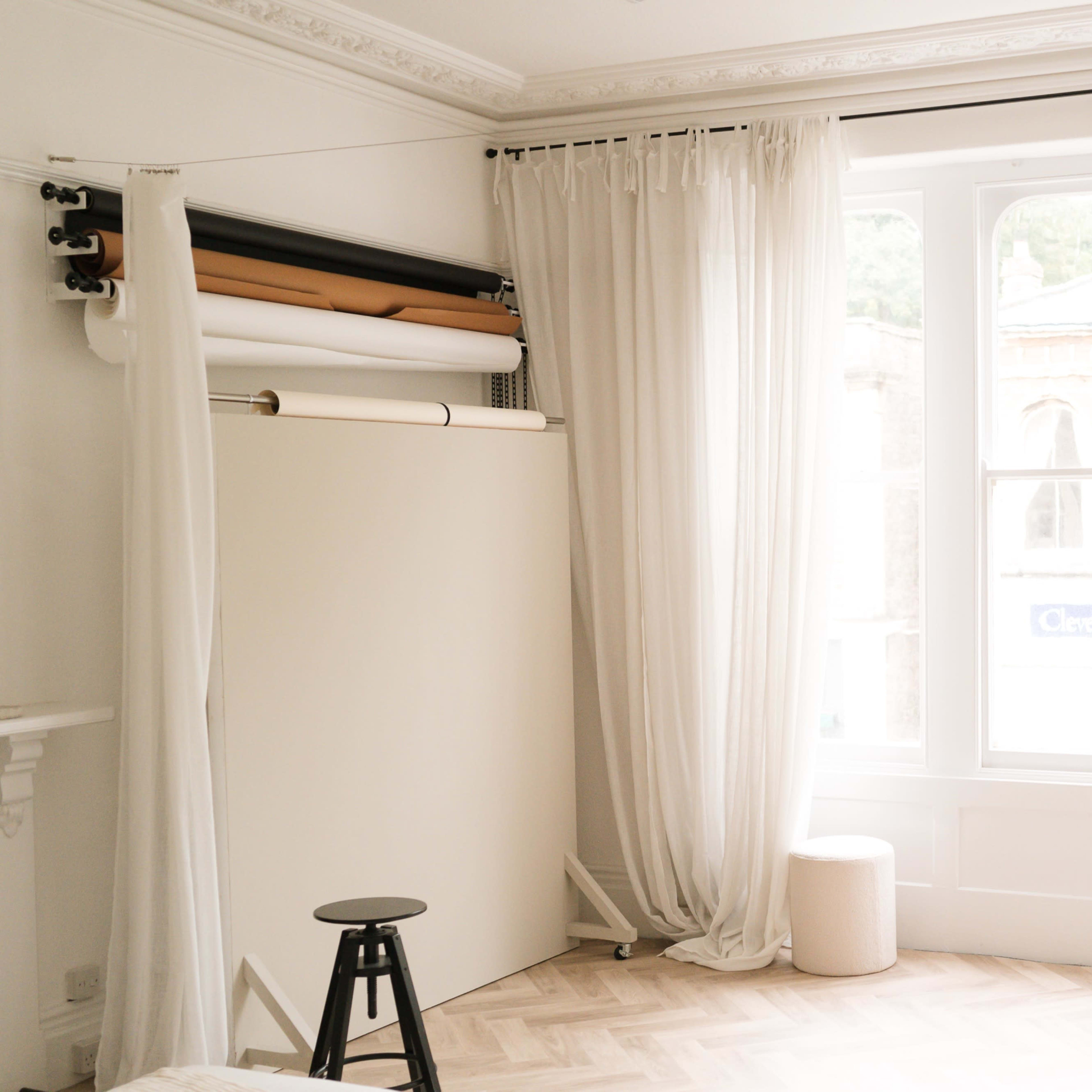 A minimalist interior with a white backdrop, a stool, and large windows adorned with sheer curtains.