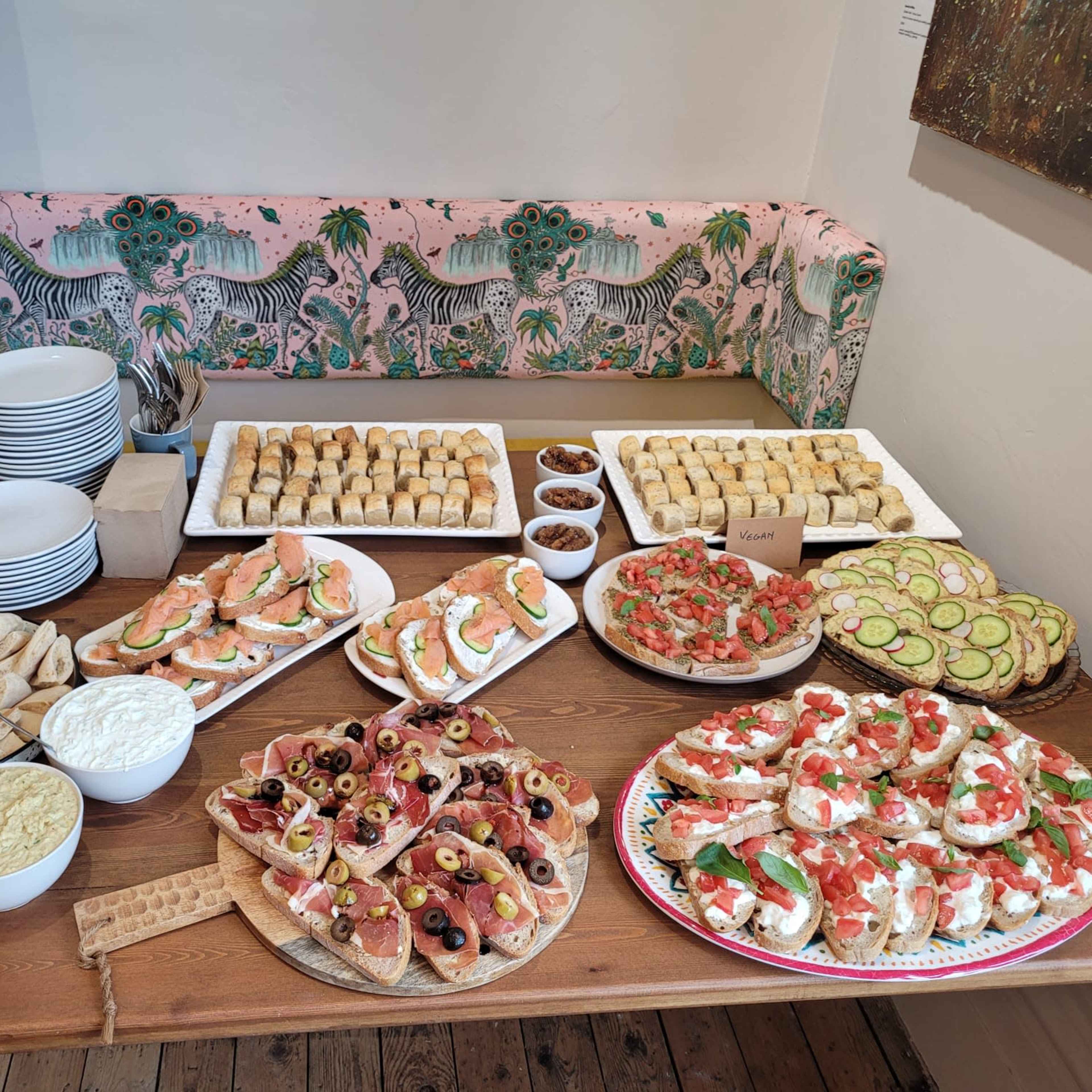 A buffet table is spread with various dishes, including platters of bruschetta, sandwiches, pastries, and dips, alongside plates and utensils for serving.