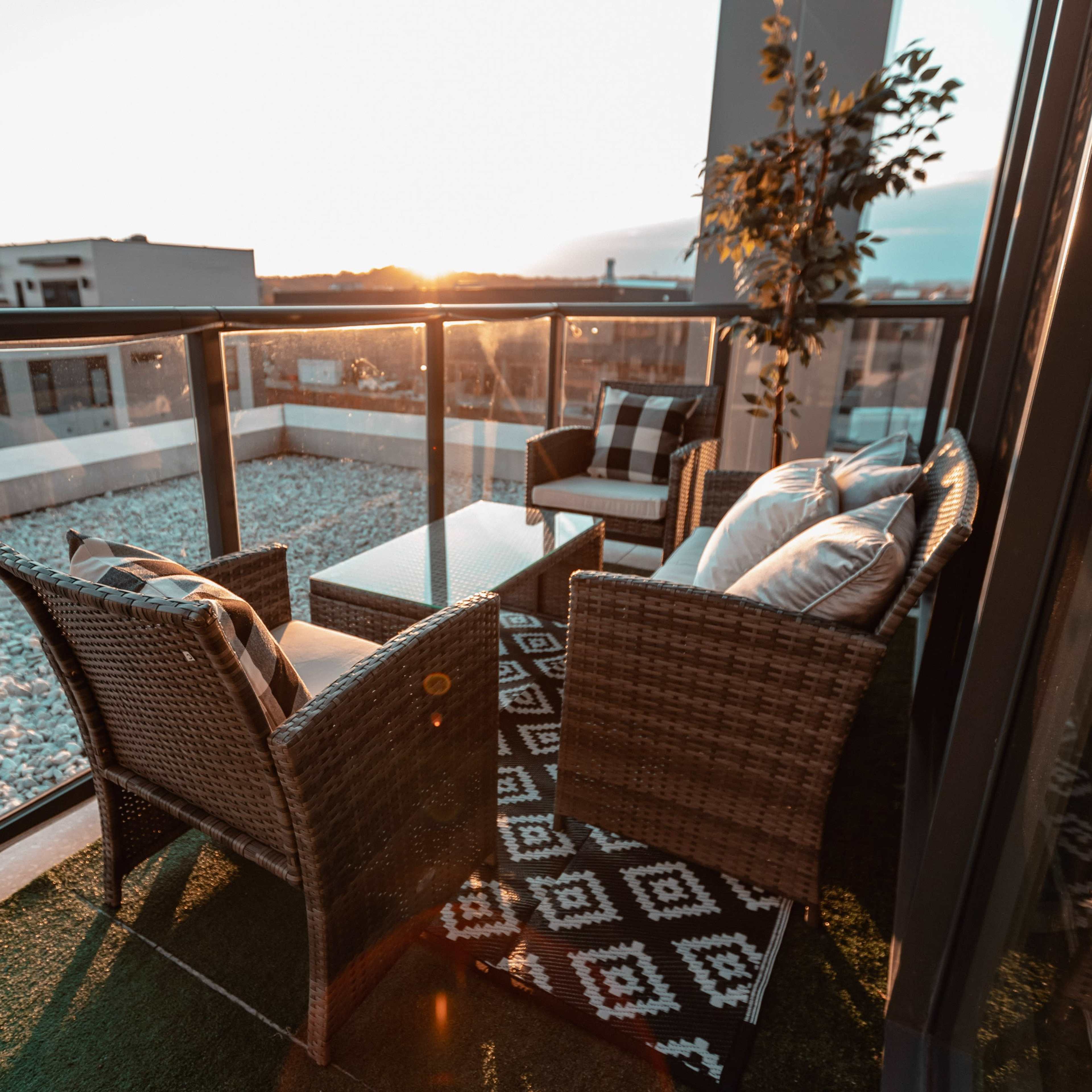 A balcony furnished with two wicker chairs, a small table, and a rug, overlooking a gravel-covered area with buildings in the background during sunset.