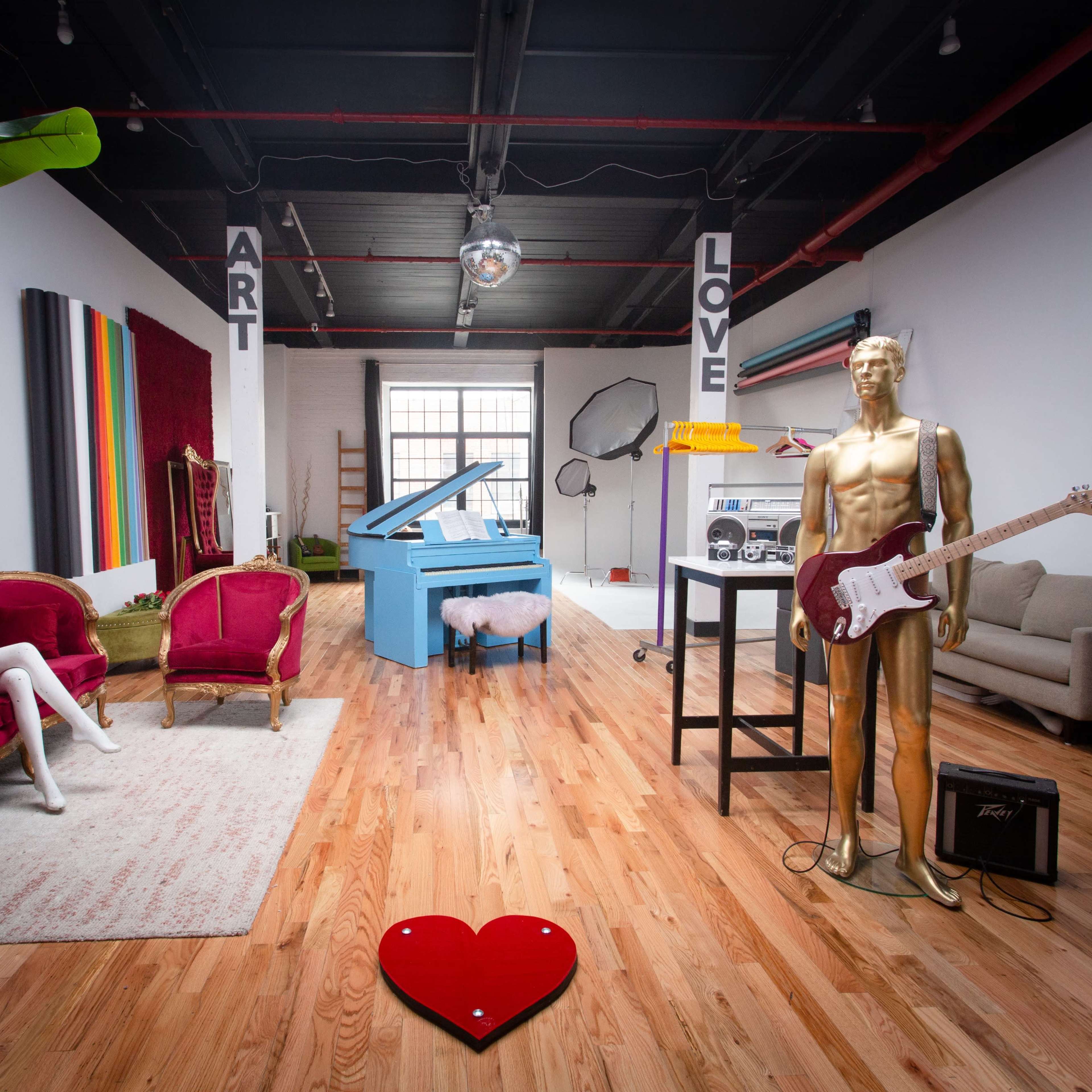 A spacious art studio featuring a blue piano, gold mannequin with a guitar, plush seating, and decorative elements like a heart-shaped design on the floor.