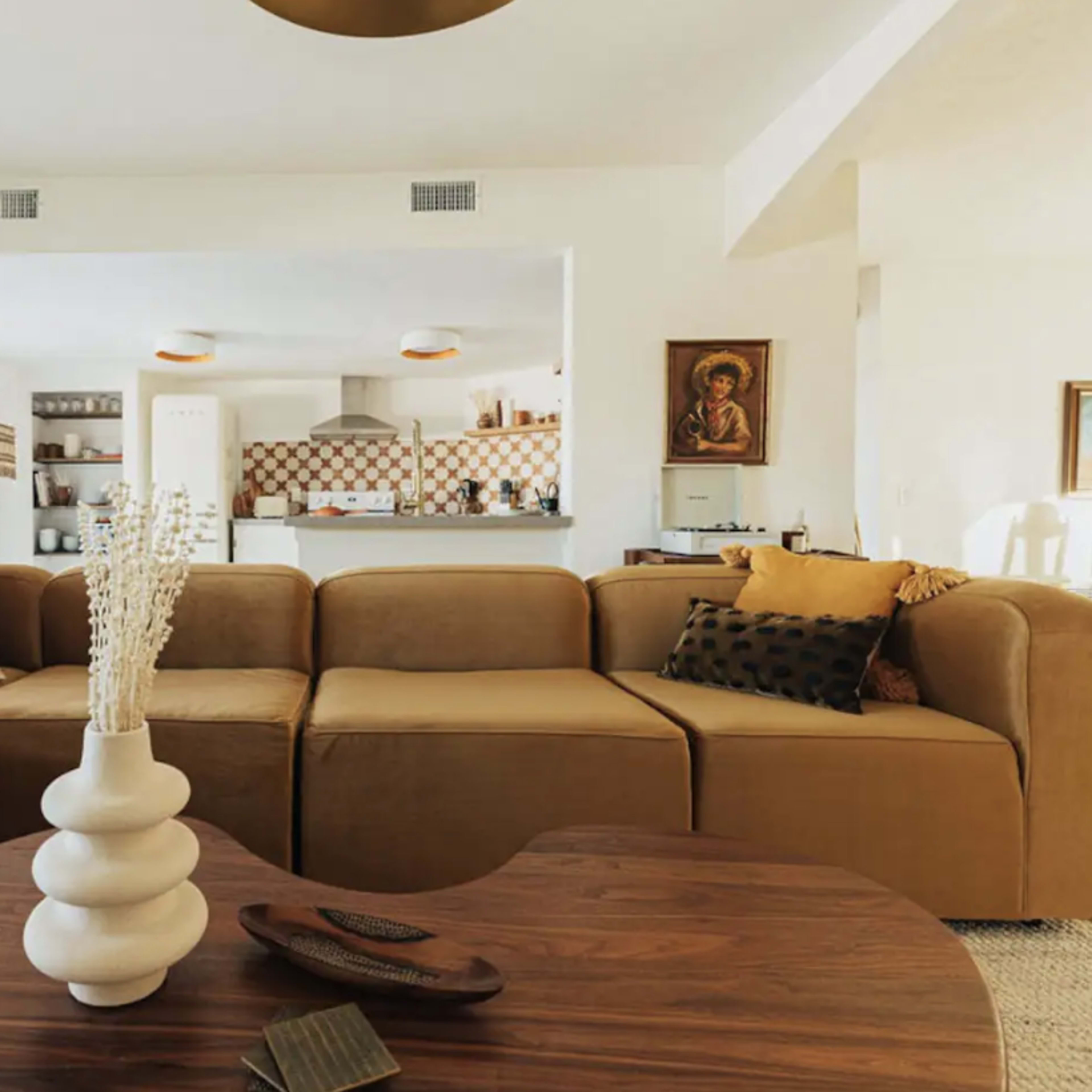 The living room features a large, L-shaped brown sectional sofa and a round wooden coffee table with decorative items on it, set against a backdrop of a kitchen area.