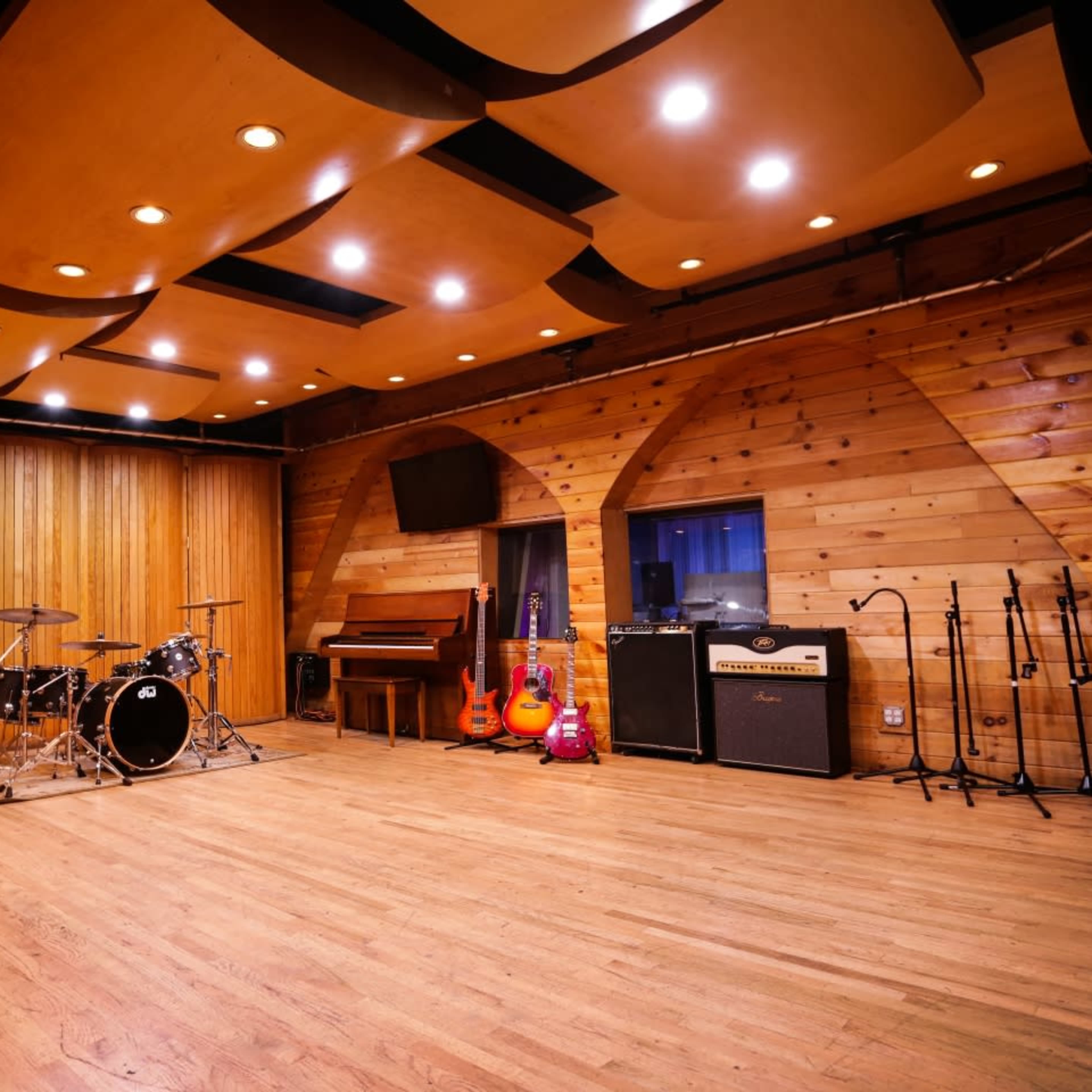 A music rehearsal space features wooden walls, a drum set, a piano, and various guitars arranged beside amplifiers.