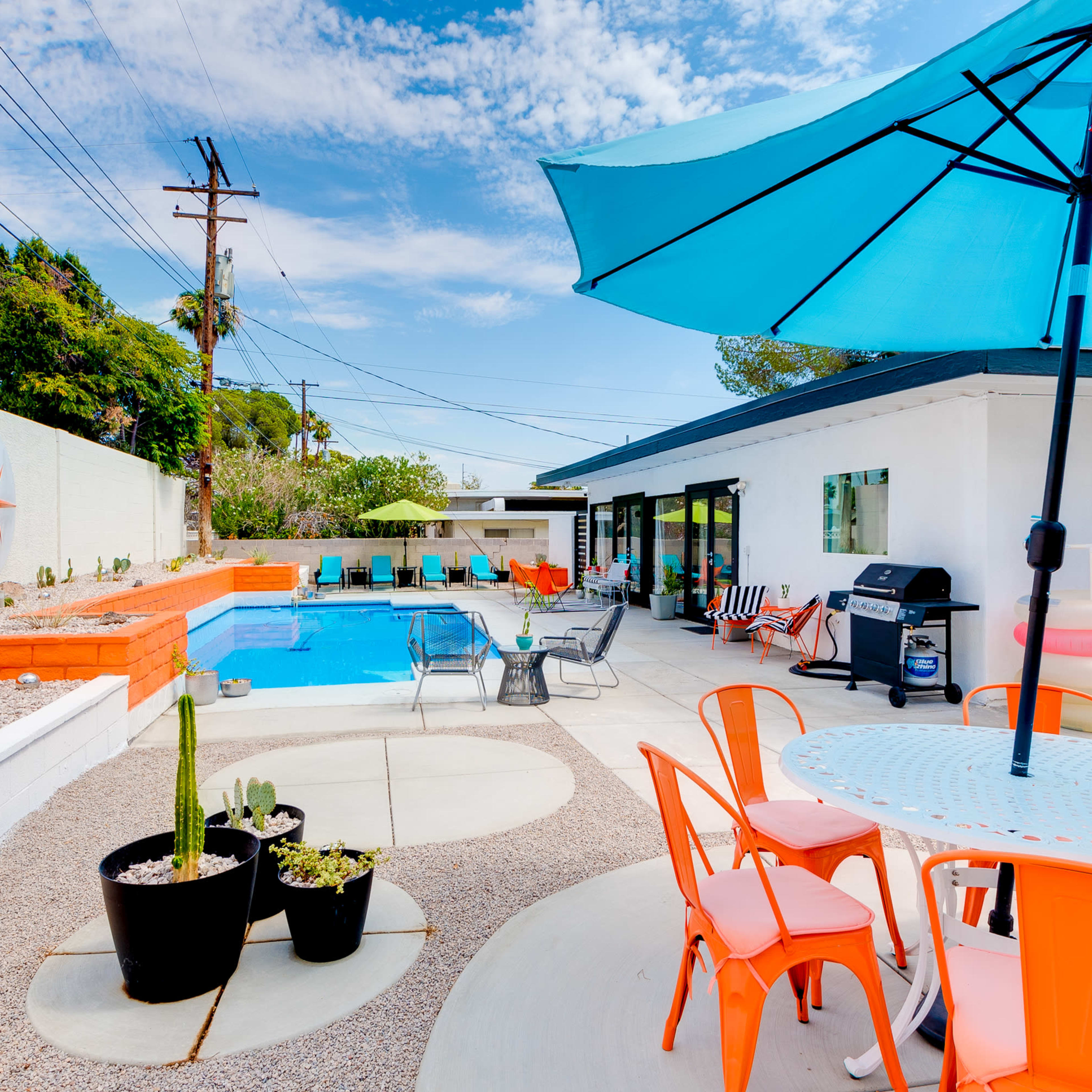 A modern outdoor space featuring a swimming pool surrounded by a patio with colorful lounge chairs, an umbrella, and a dining table.