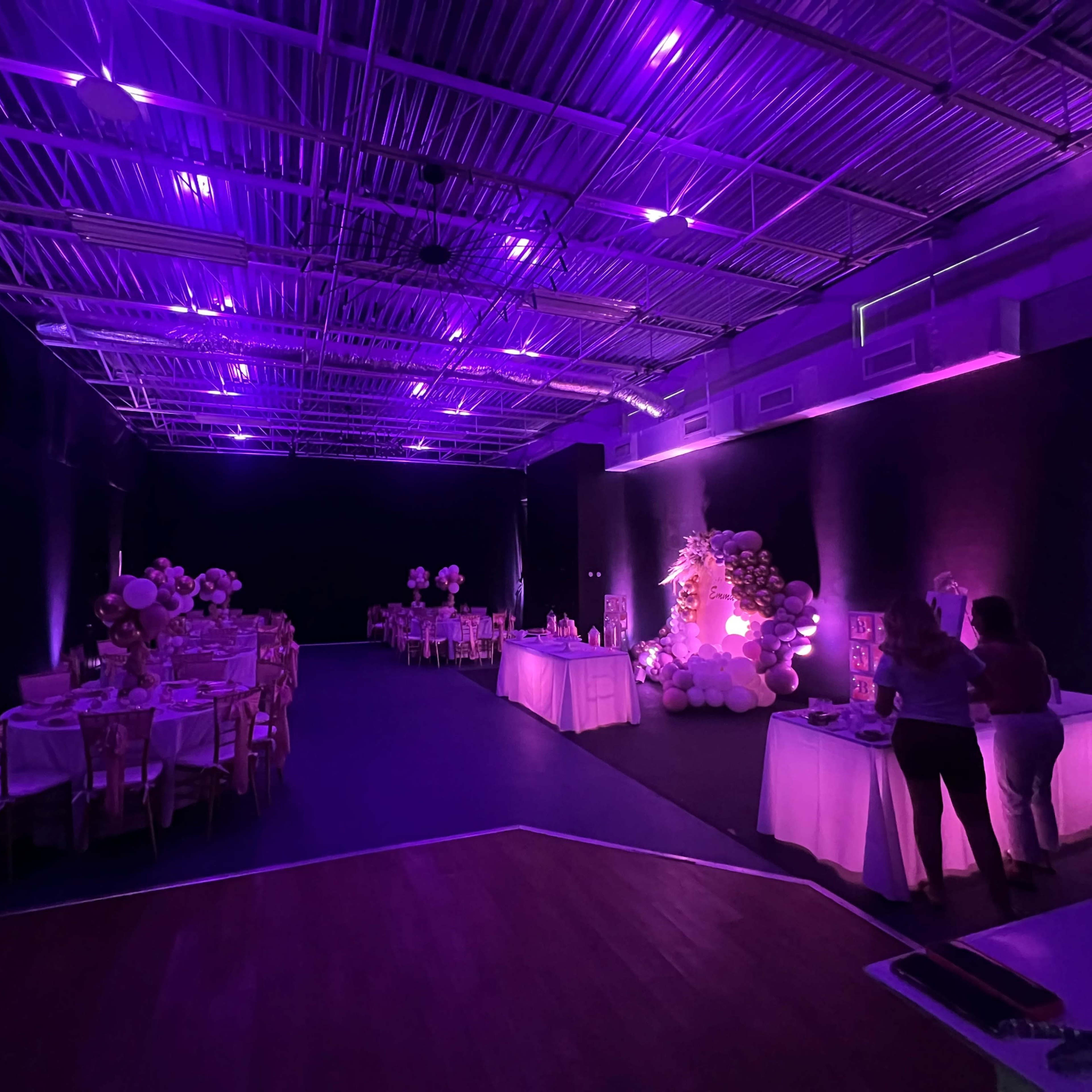 A dimly lit event space decorated with purple lighting, featuring tables set for dining and a balloon display.