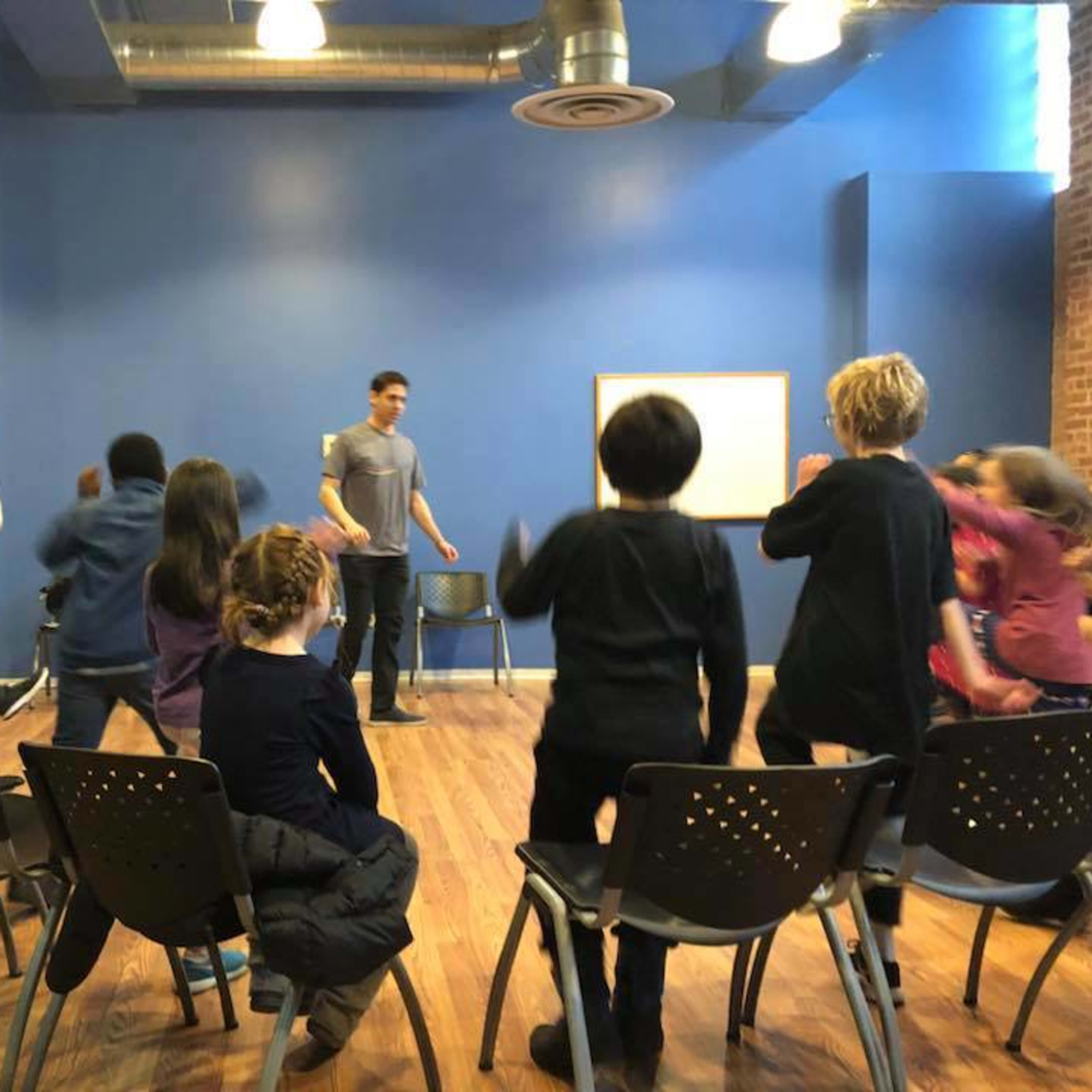 A group of children participates in a lively activity while seated in chairs facing an adult in a blue room.