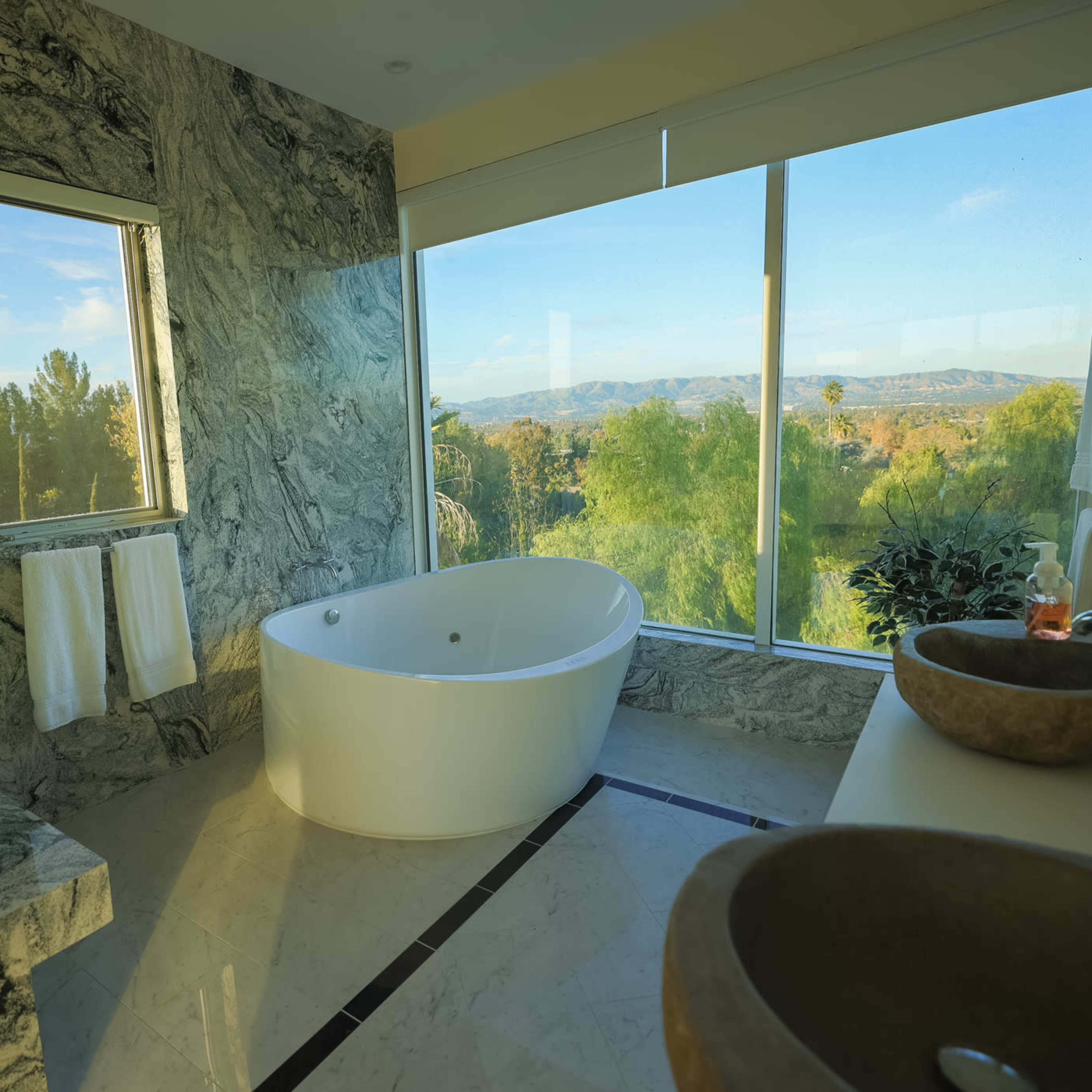 The bathroom features a freestanding white tub, two stone sinks, and large windows offering a view of the surrounding landscape.