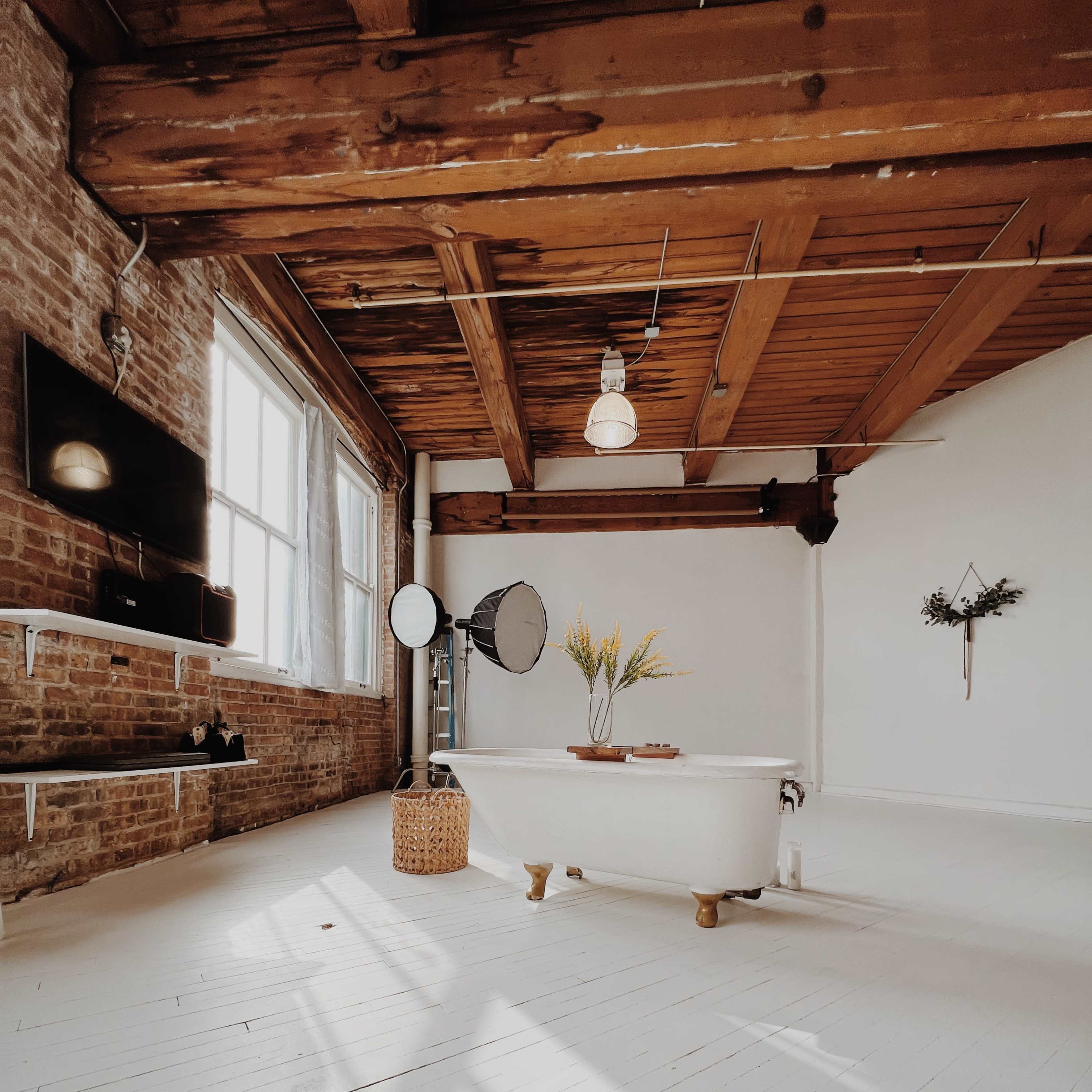 A spacious, well-lit room with exposed brick walls, wooden beams, and a vintage bathtub positioned in the center, complemented by a plant arrangement on a table.