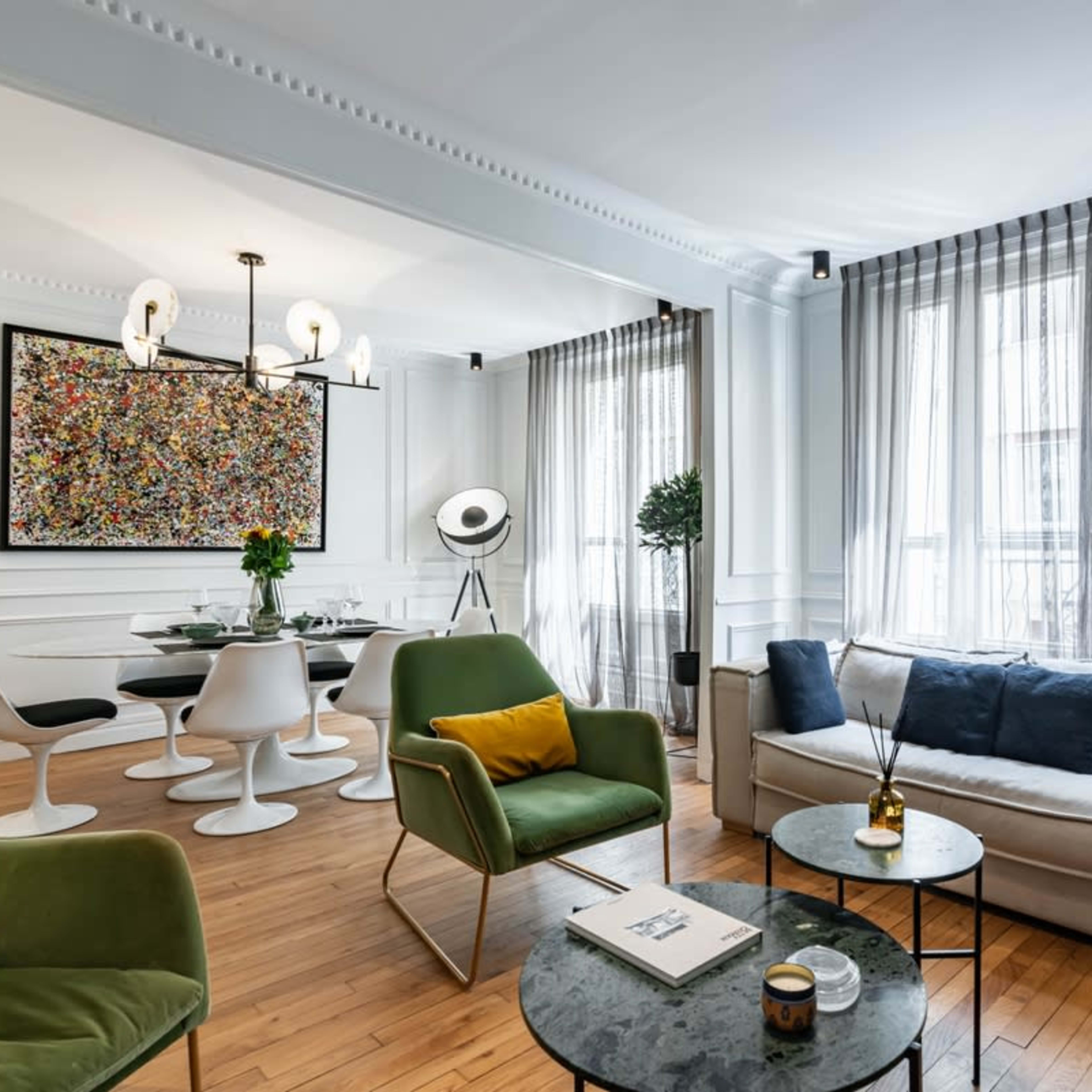 A modern living room with a mix of green and white furniture, a round table with white chairs, and large windows draped with sheer curtains.