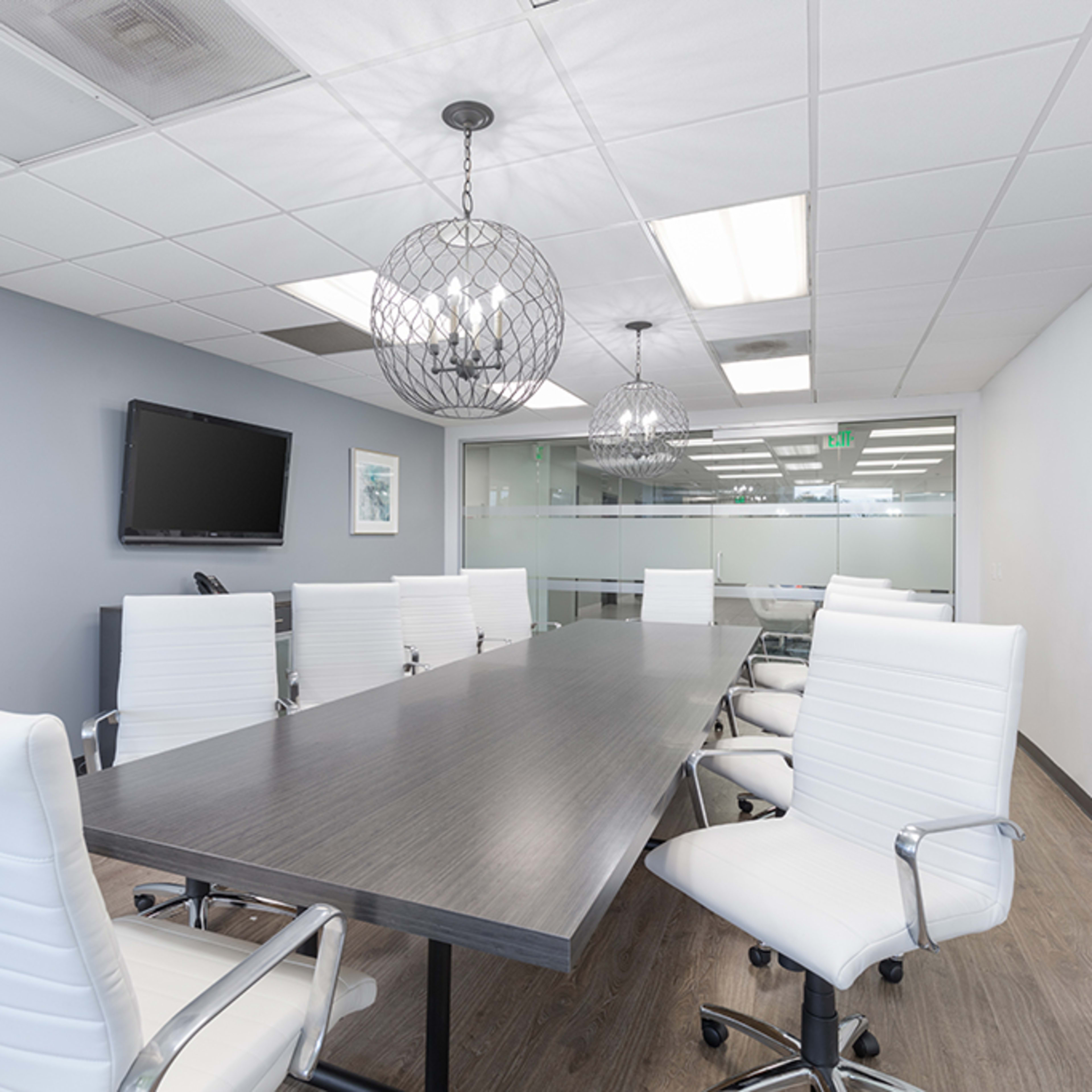 A modern conference room features a long wooden table surrounded by white ergonomic chairs, with pendant lights and a wall-mounted screen.
