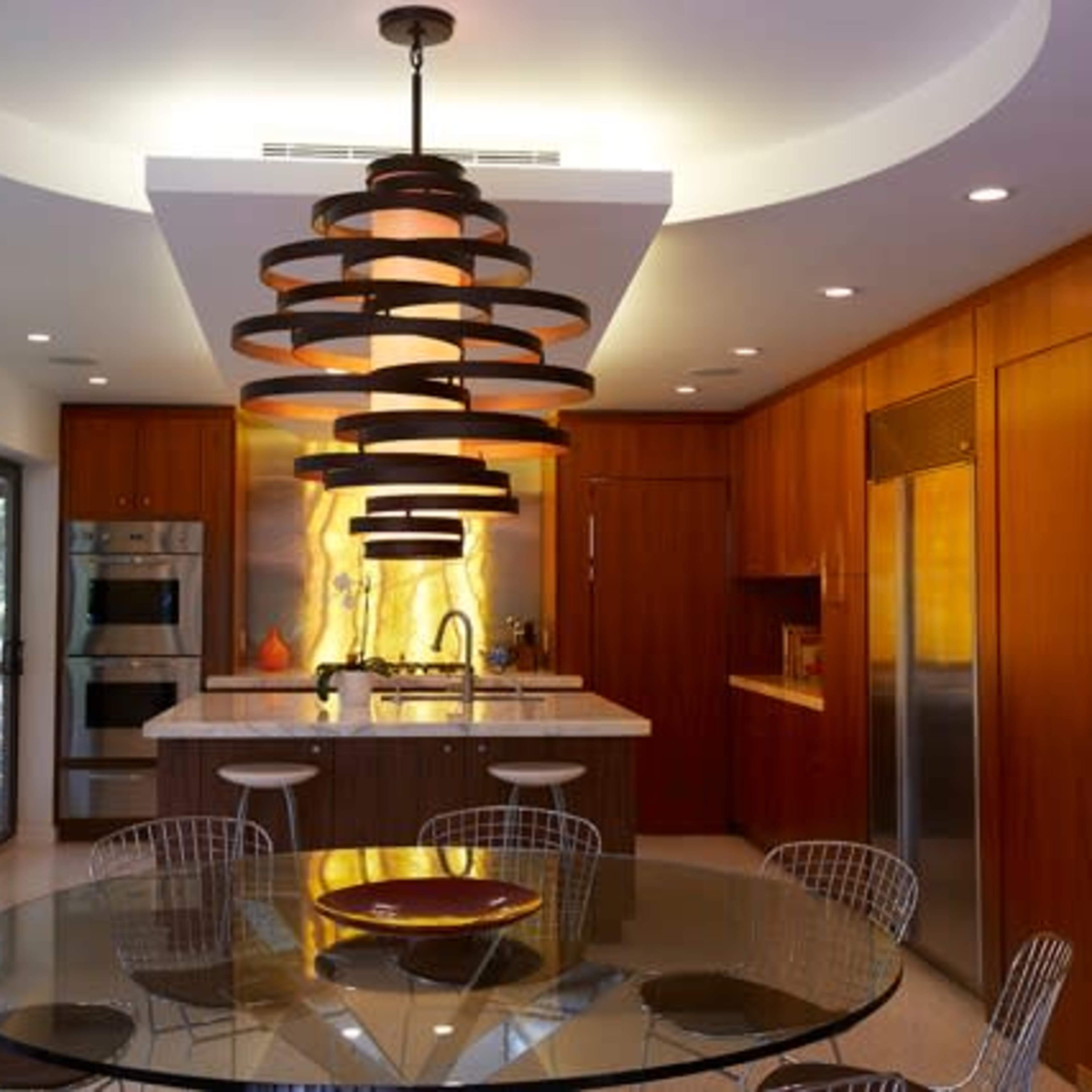 A modern kitchen features wooden cabinetry, a central island with a circular glass dining table, and a distinctive pendant light.