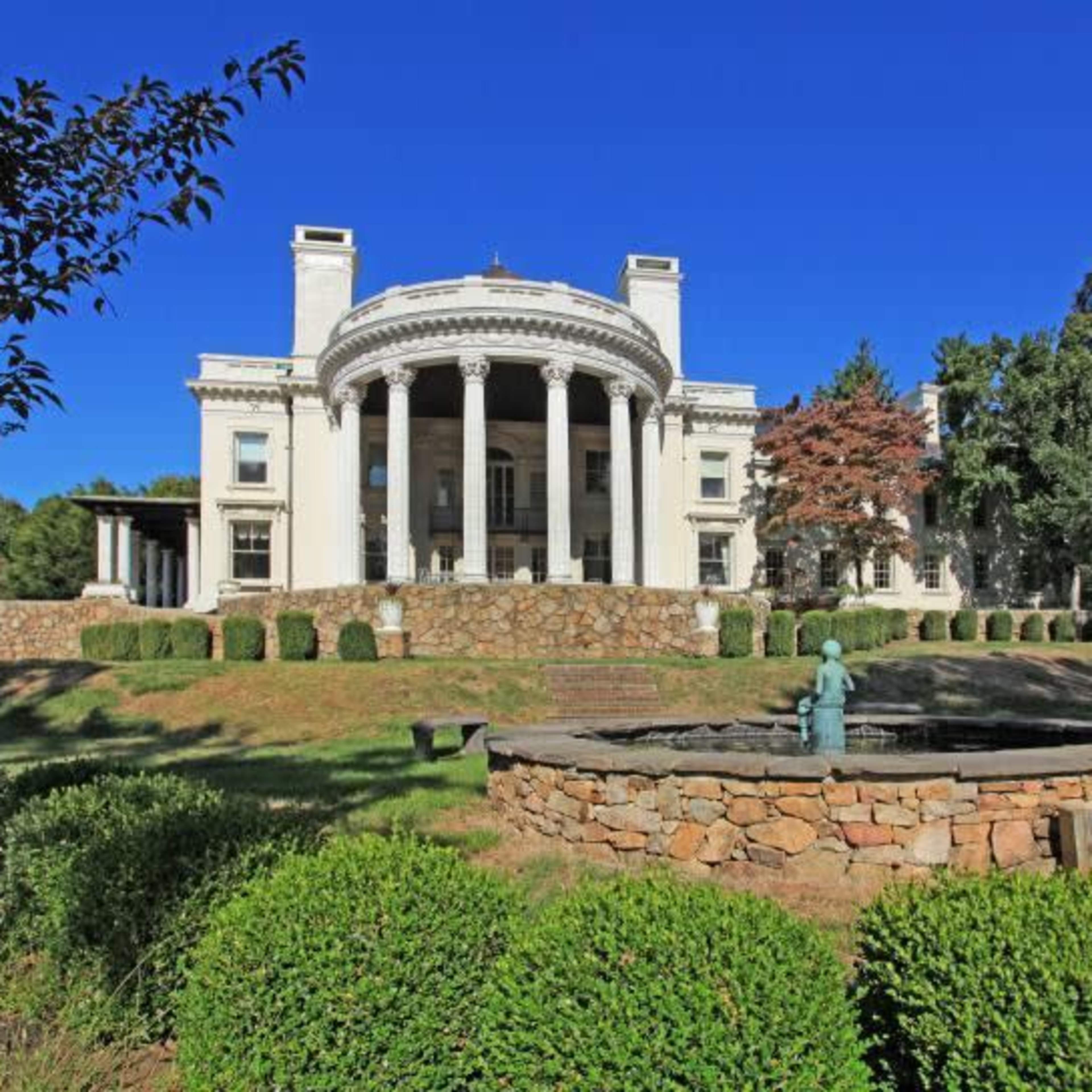 A grand mansion with a circular colonnade, set on a landscaped property featuring a stone fountain and neatly trimmed shrubs.