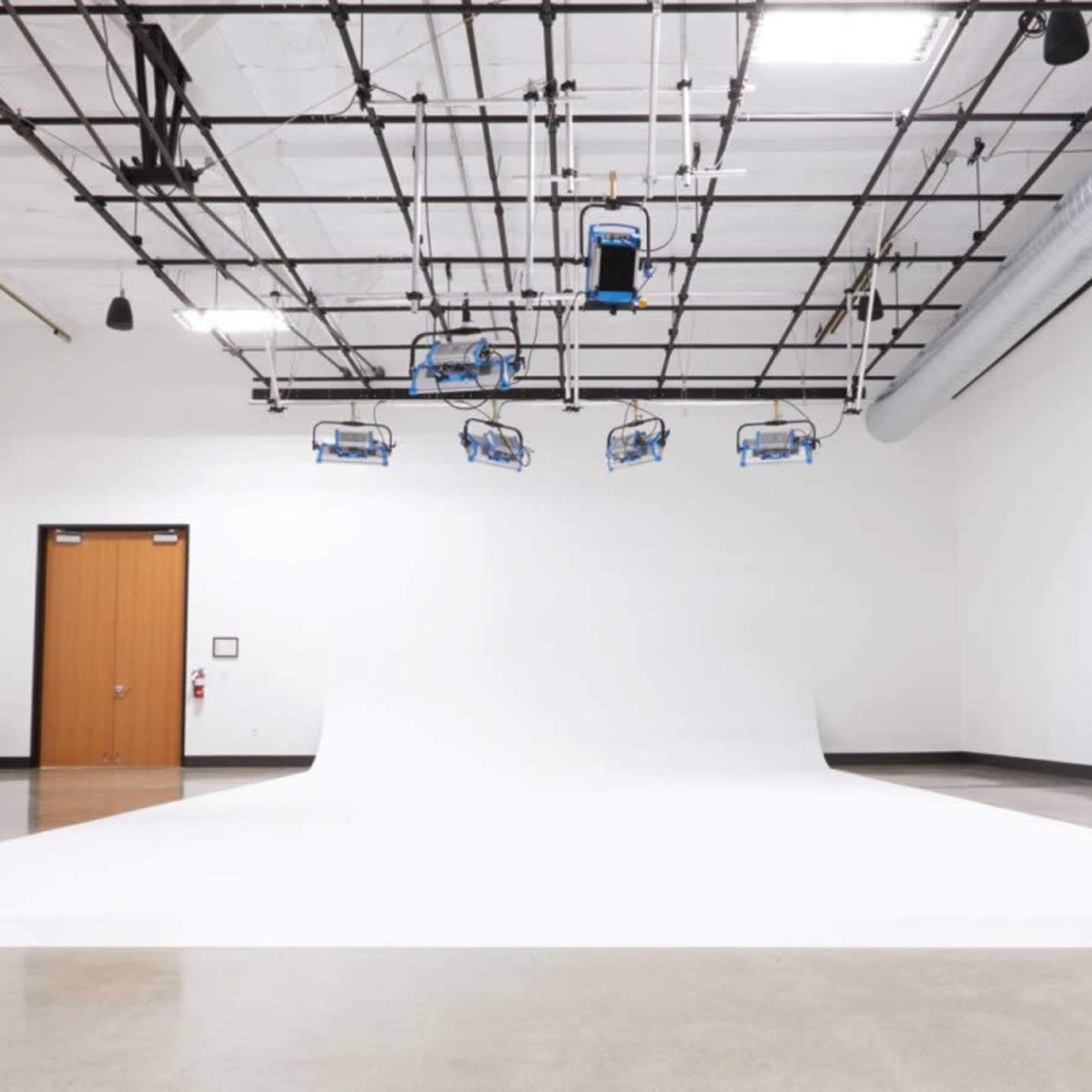 A spacious, empty studio with a white backdrop, overhead lighting fixtures, and a closed door in the background.