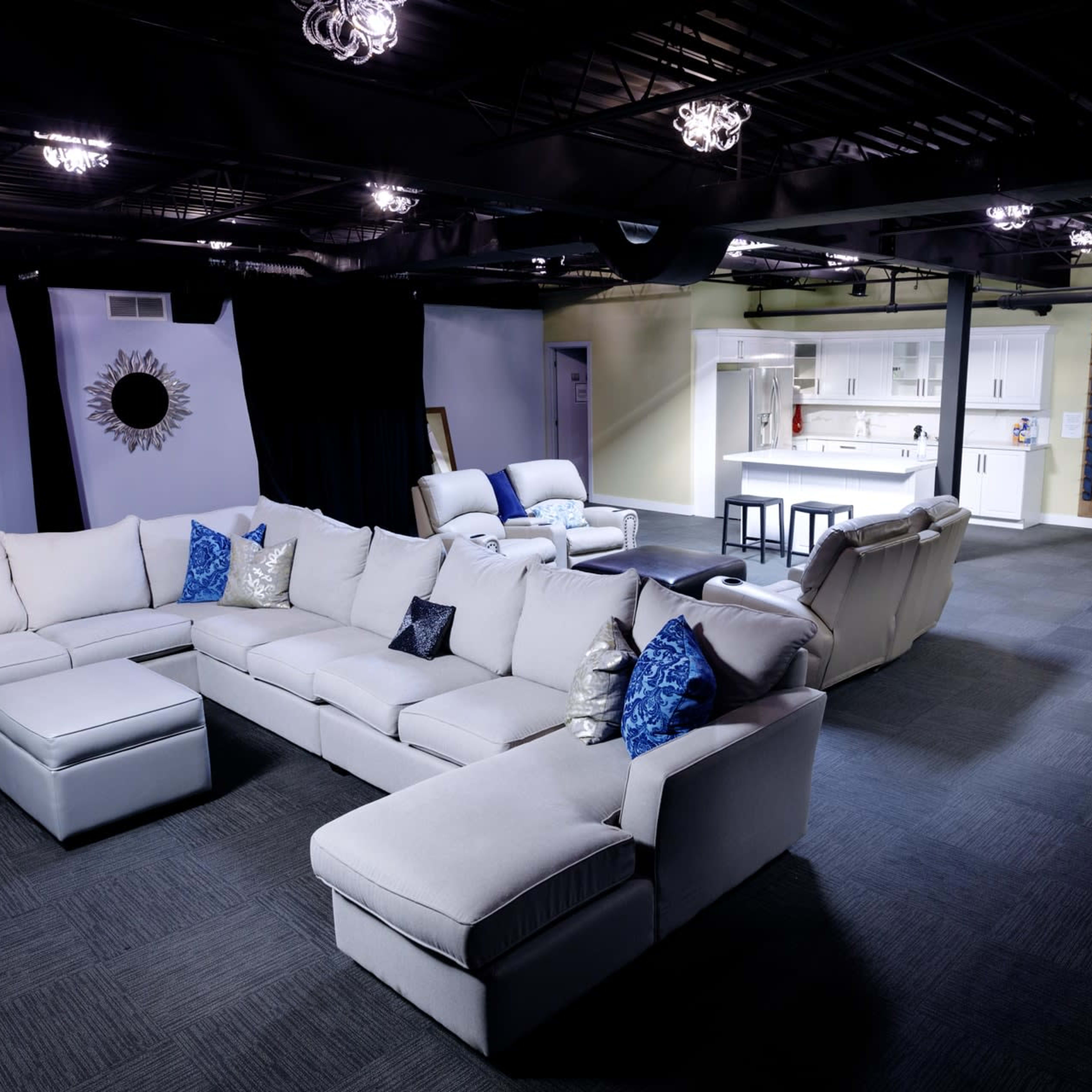 A spacious living area featuring a large sectional sofa with decorative pillows, a minimalist dining set, and a separate kitchen area with white cabinets in the background.