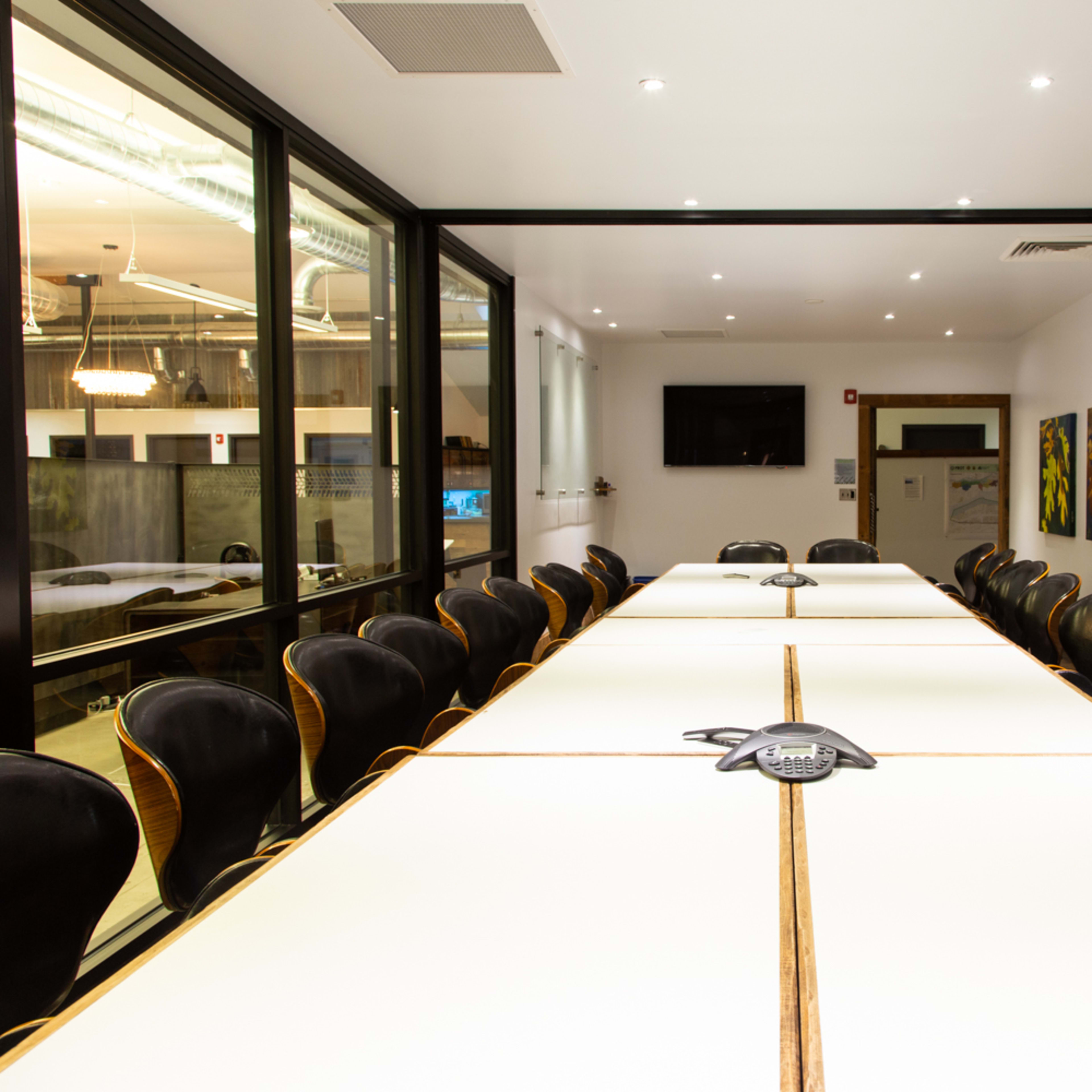 A large conference room features a long table with multiple chairs arranged around it, equipped with a speakerphone and wall-mounted displays.