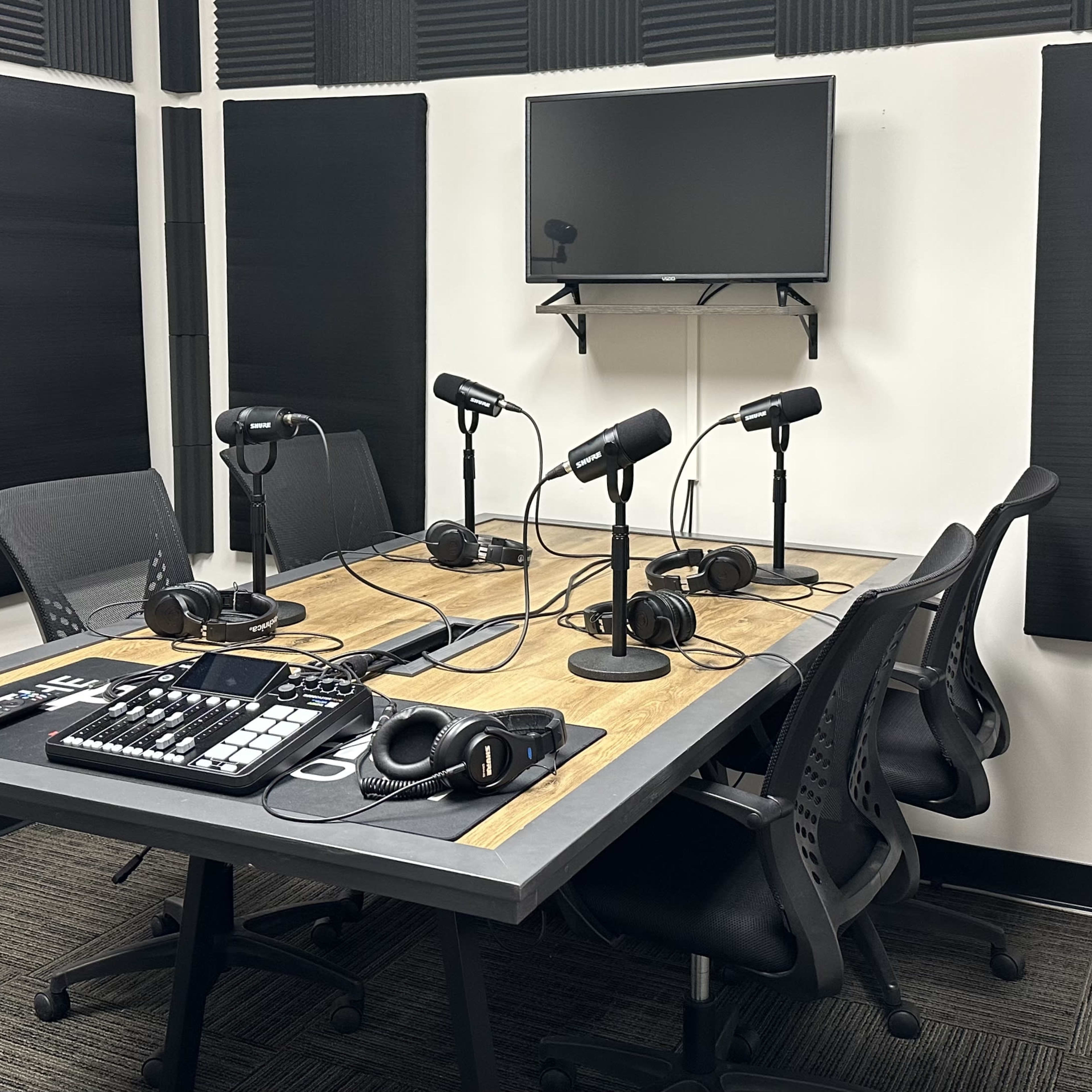 A soundproof recording studio with a rectangular table equipped with four microphones, chairs, a computer interface, and sound-absorbing panels on the walls.