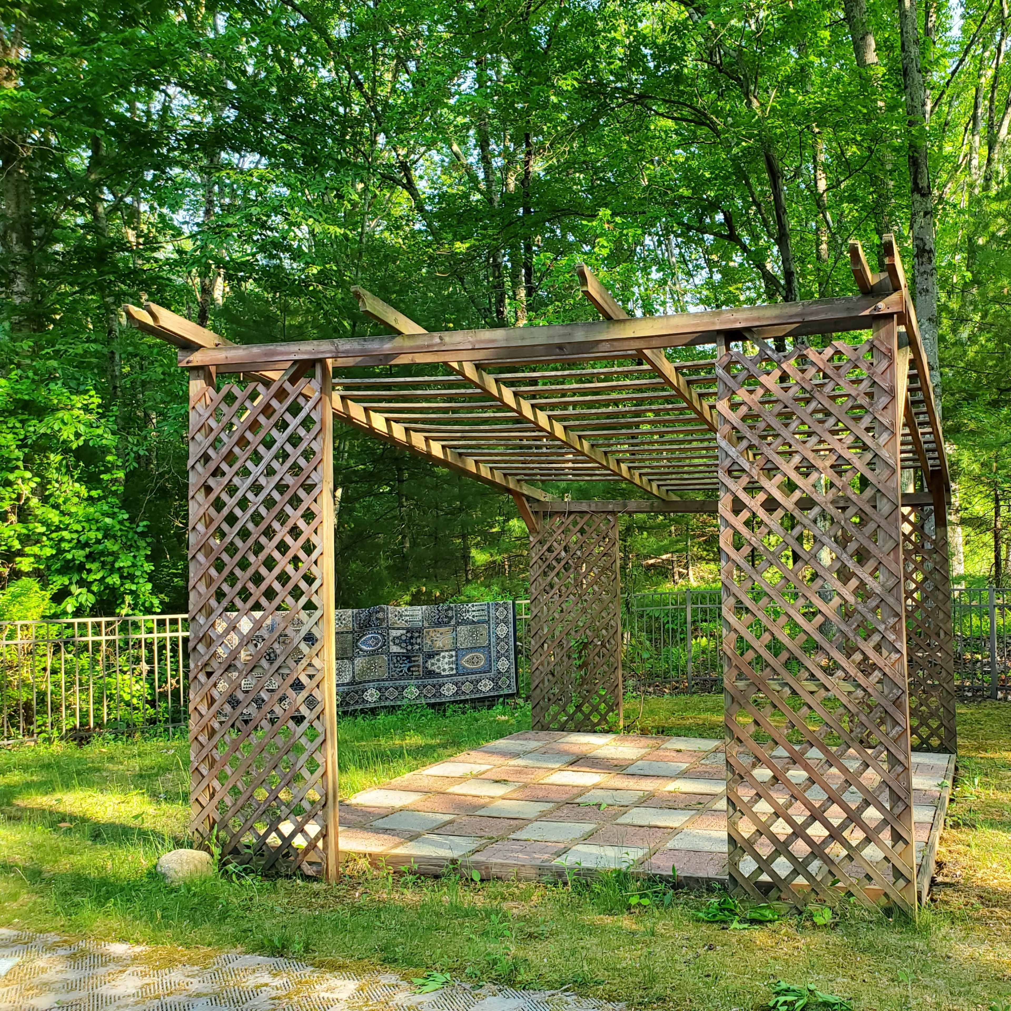 A wooden lattice gazebo stands in a grassy area surrounded by trees, with a patterned blanket draped on one side.