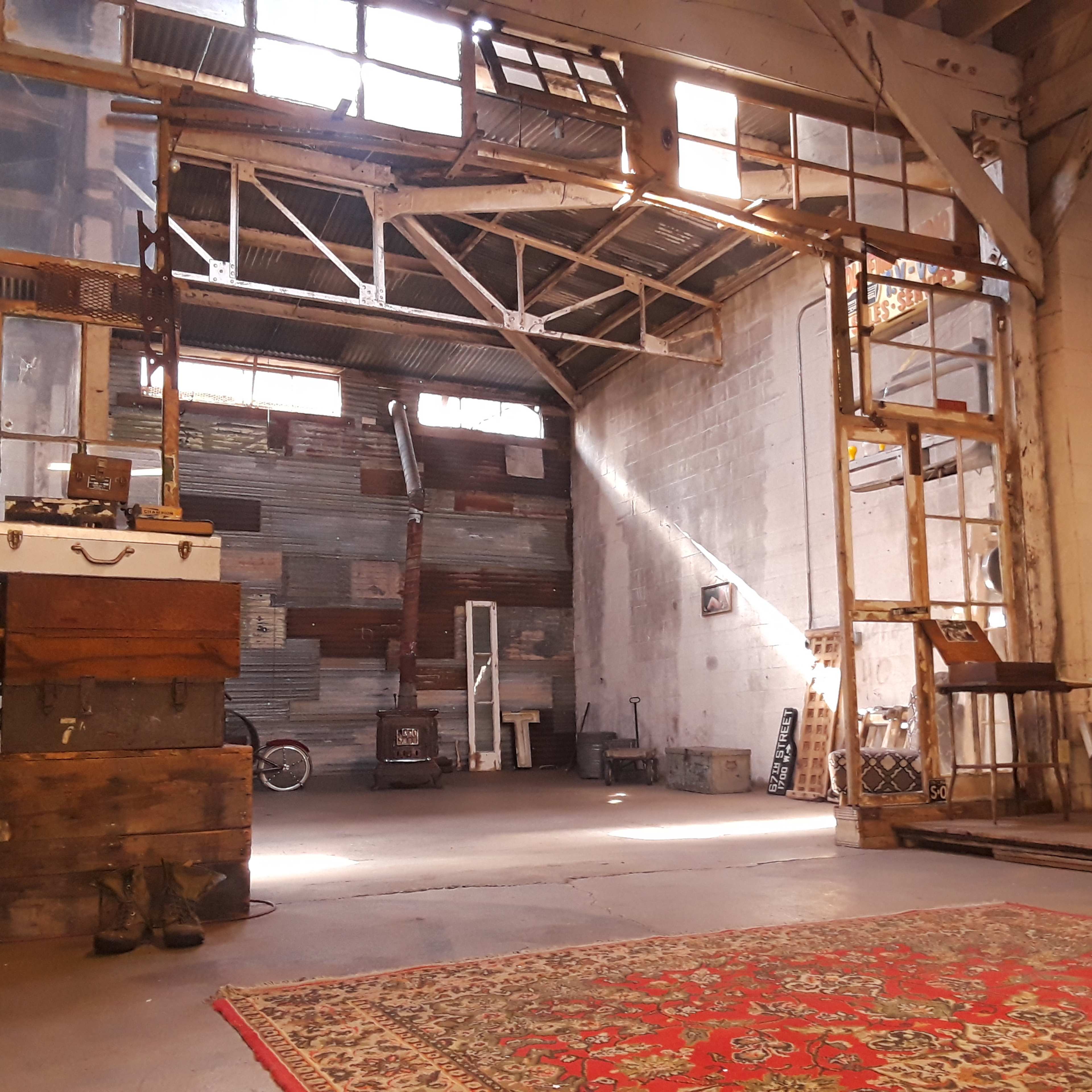 An industrial space with exposed beams, wooden crates, and a patterned rug on the floor, illuminated by natural light coming through large windows.