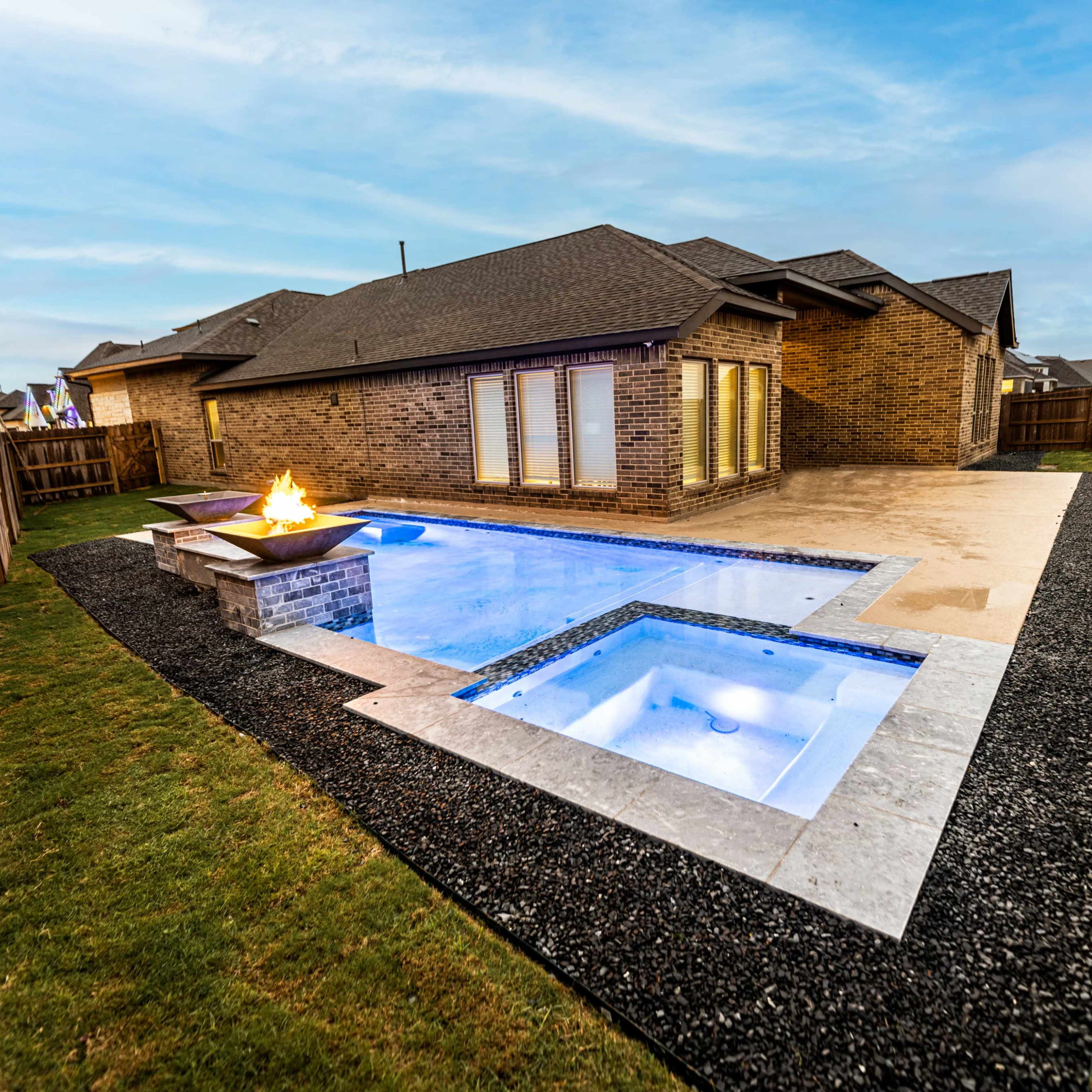 A backyard with a swimming pool, a hot tub, and a fire pit surrounded by stone and grass, enclosed by wooden fencing.