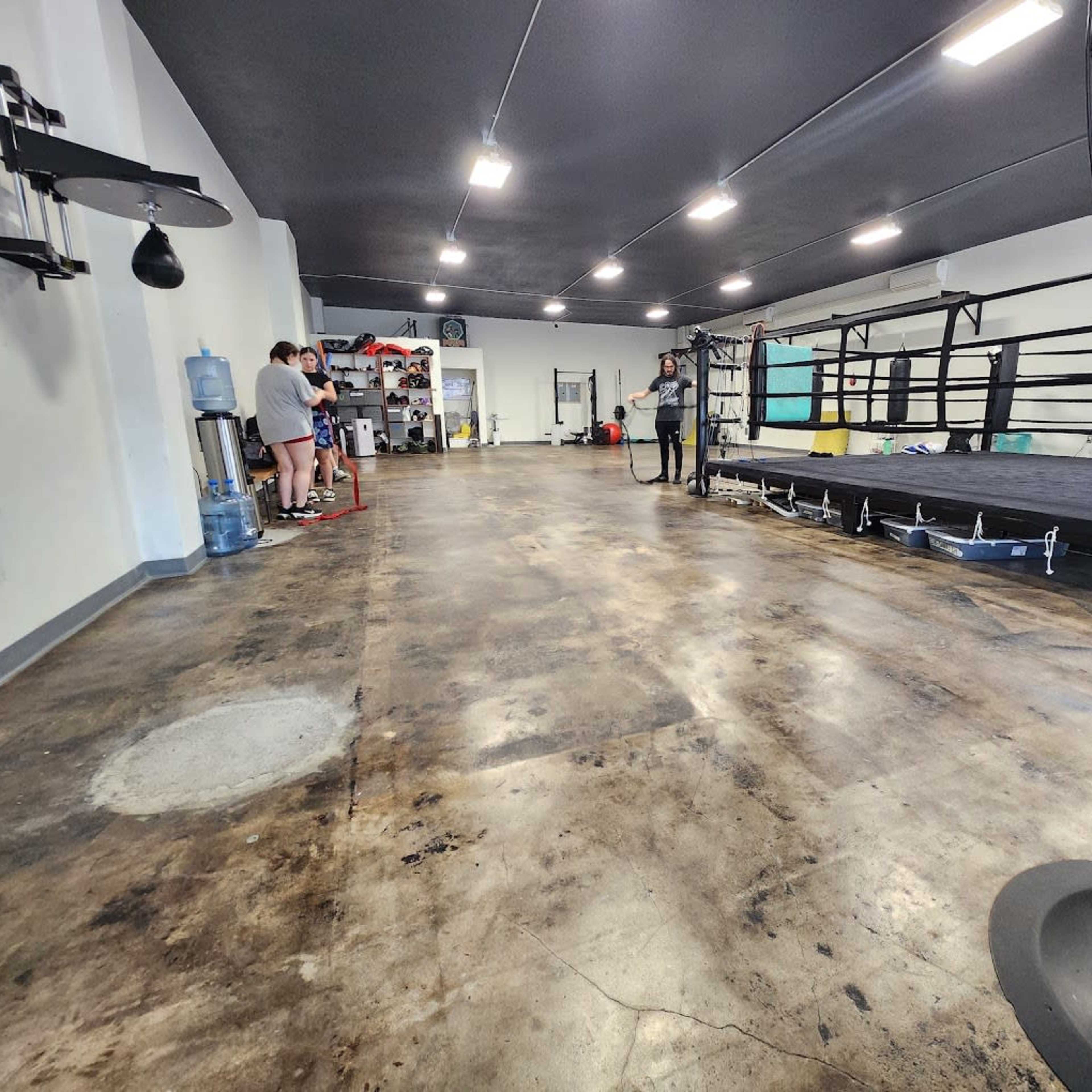 A spacious indoor boxing gym with a ring in the center, workout equipment along the walls, and a few people standing near a water cooler.