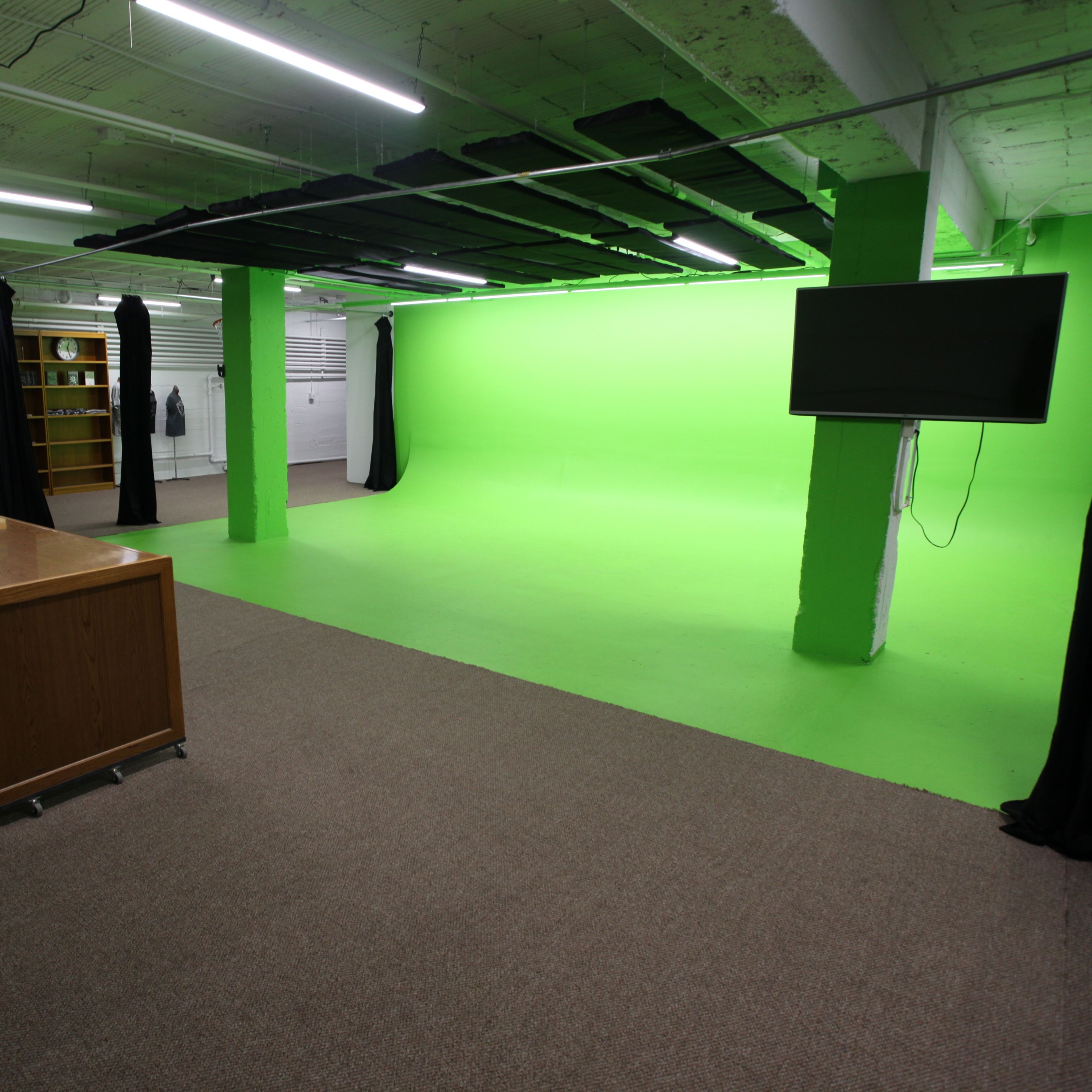 An indoor studio space featuring a green screen backdrop, a wooden desk with a chair, and shelves filled with various items.