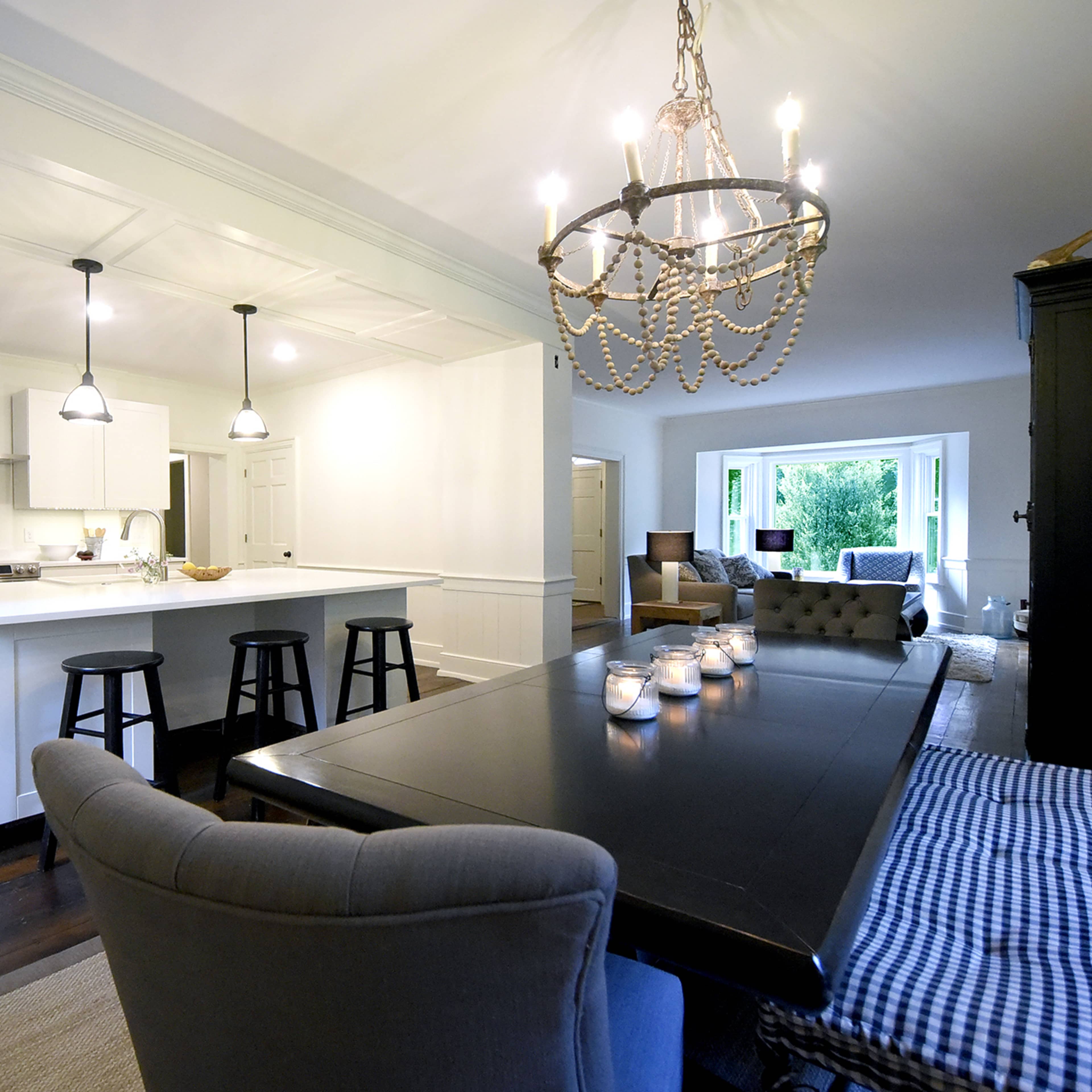 A modern kitchen with a large dining table, black stools at the island, and a chandelier overhead, set in a bright room with wood flooring.
