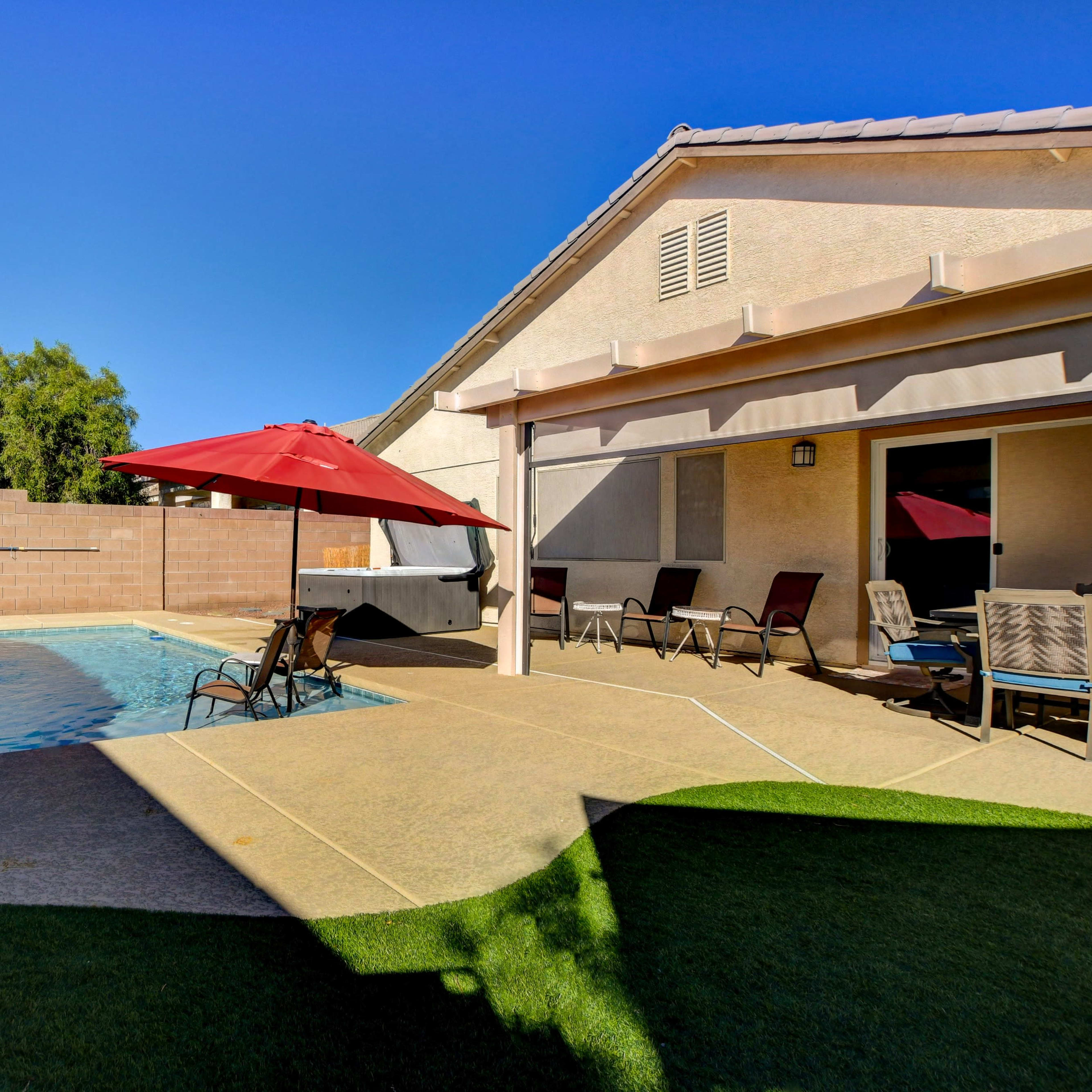 A backyard with a swimming pool, a red umbrella, lounge chairs, and a beige house under a clear blue sky.