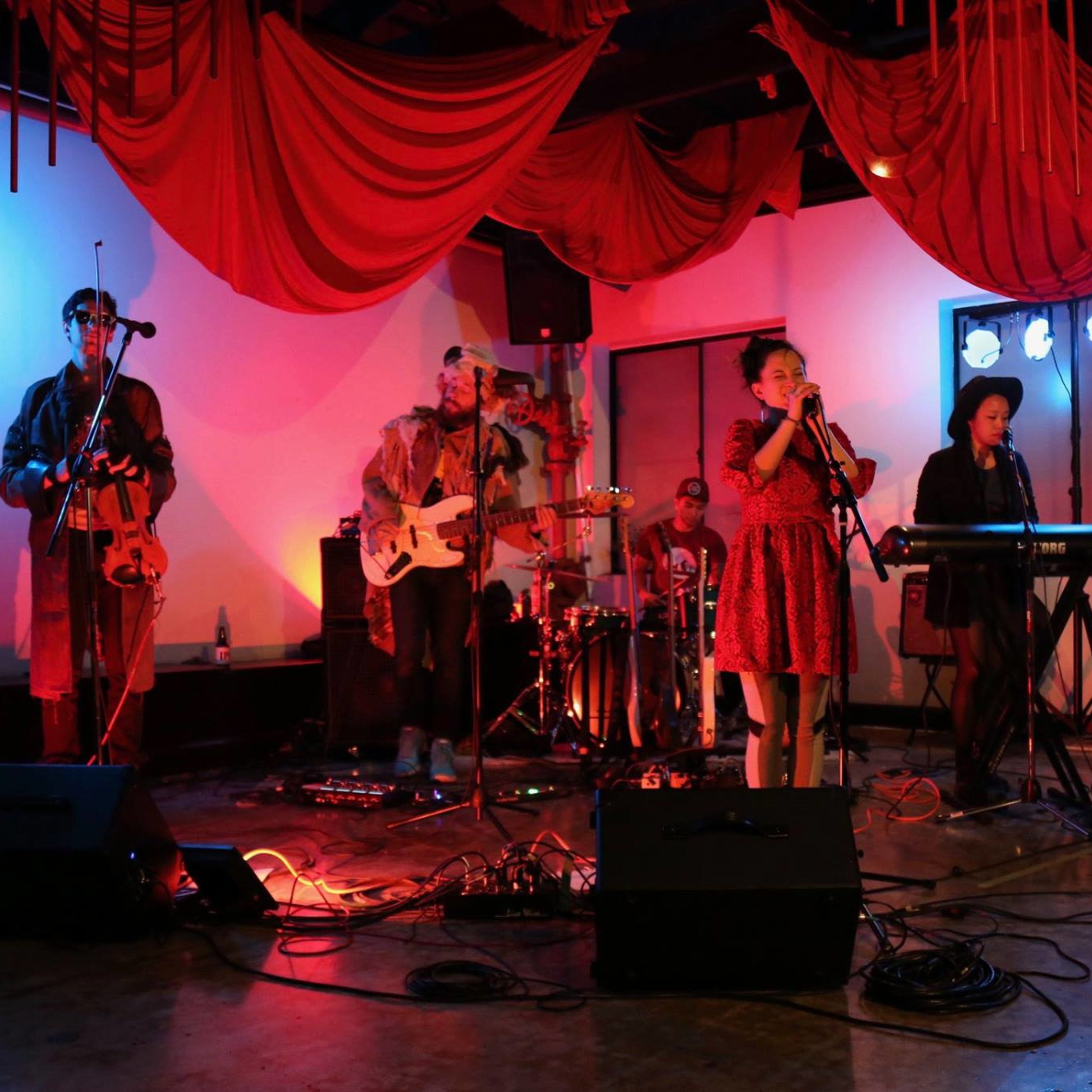 A musical band performs on stage in a dimly lit venue with red fabric decor and various instruments set up around them.