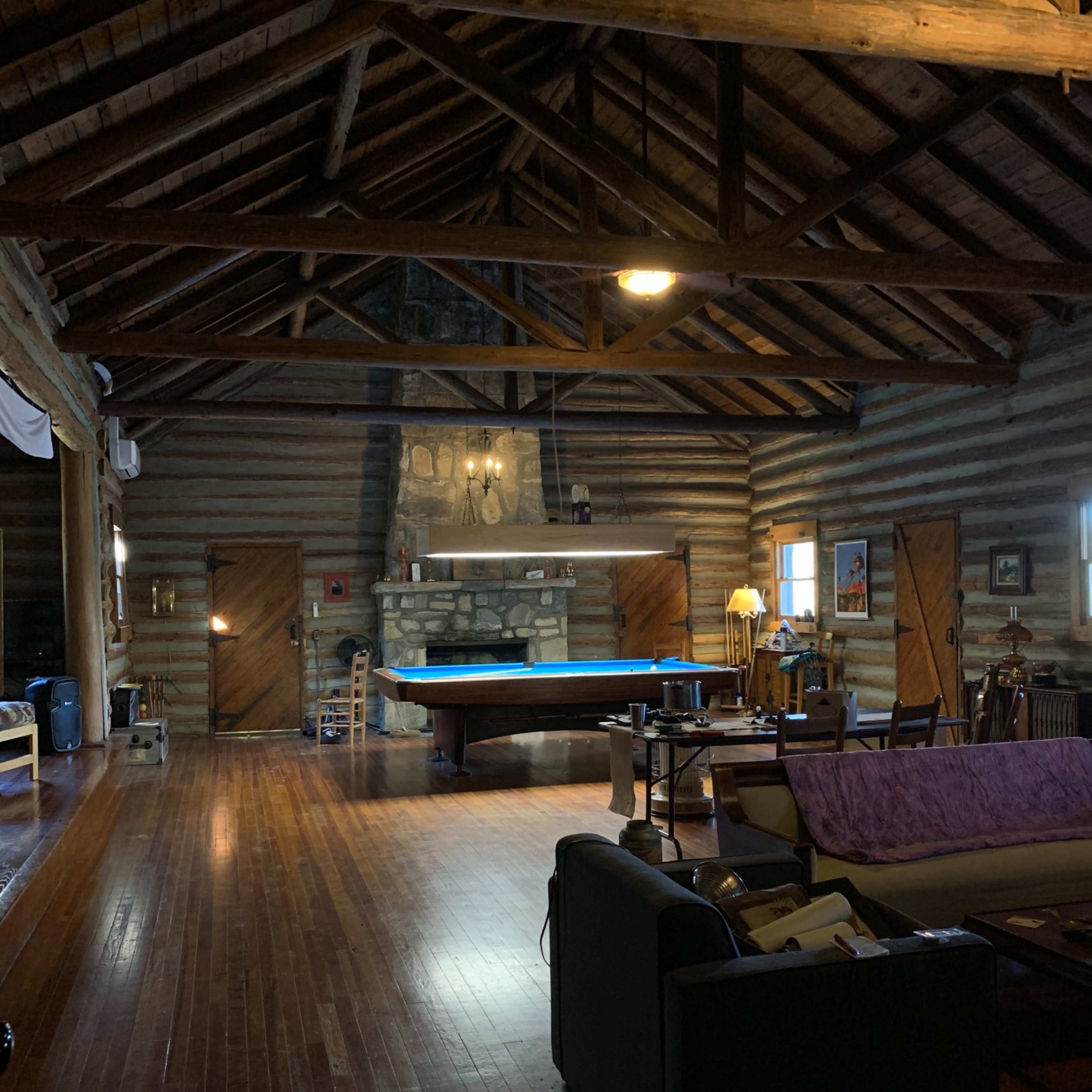 A spacious cabin interior with wooden beams and a pool table in the center, surrounded by various furniture and a stone fireplace.