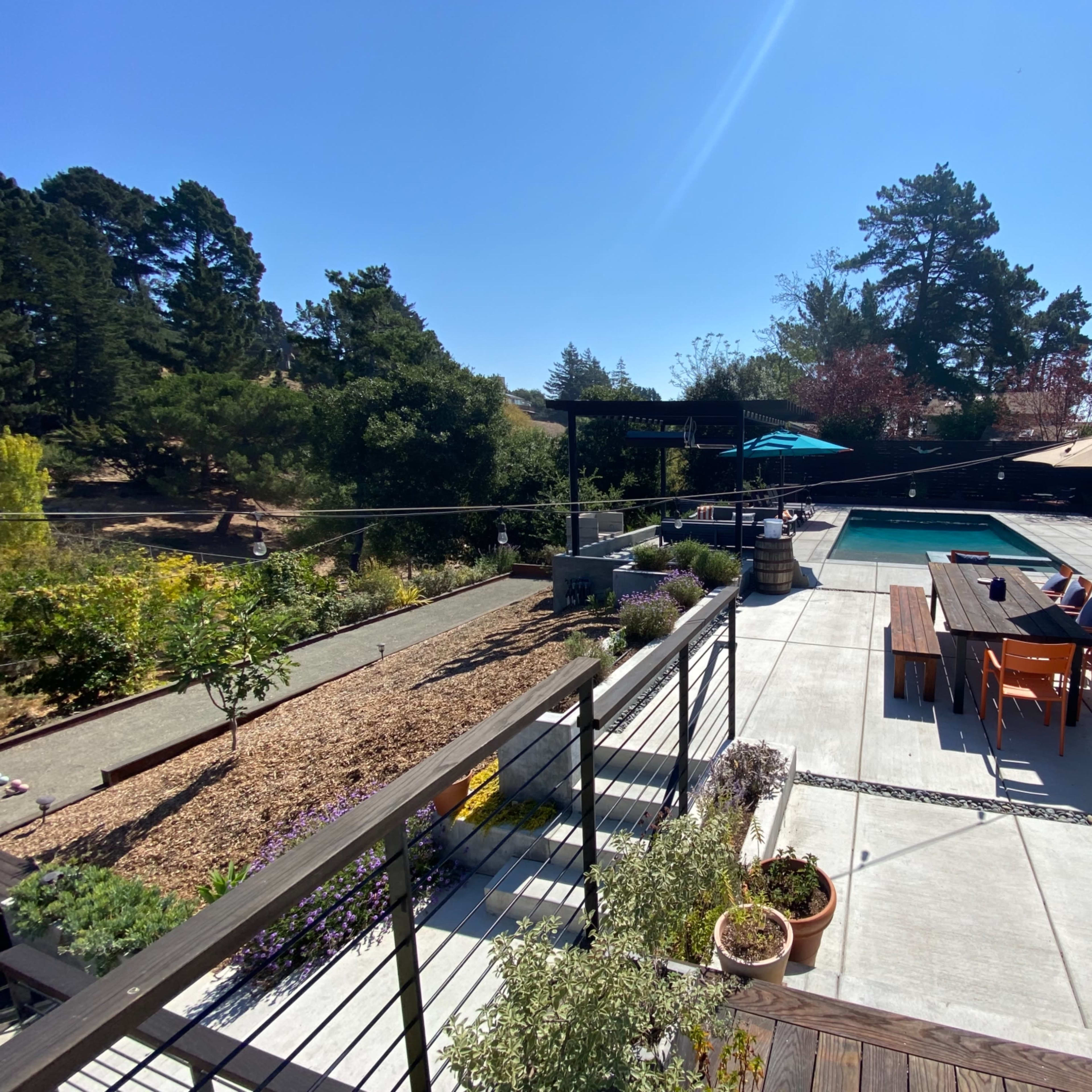 A backyard with a swimming pool, a dining table, and a barbeque grill, surrounded by landscaping and trees under a clear blue sky.