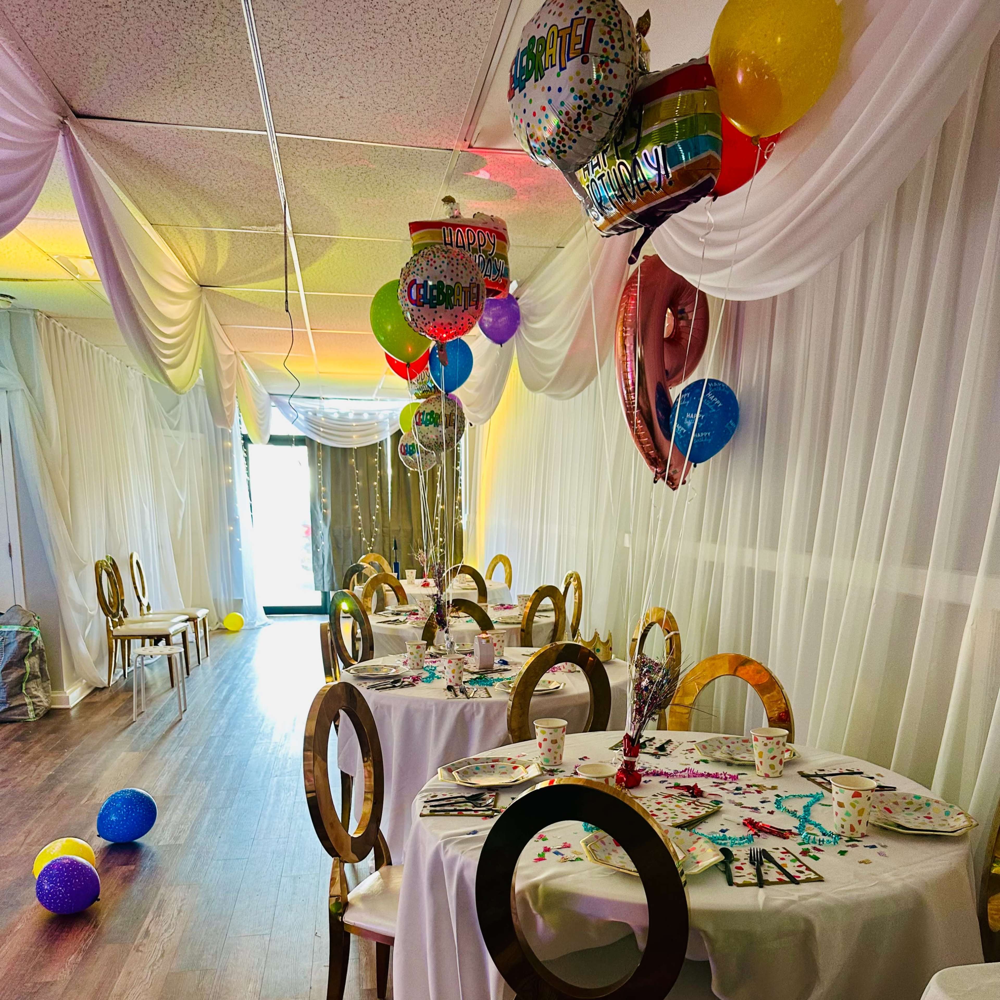 A decorated event space with round tables covered in white tablecloths, colorful balloons, and party decorations scattered on the floor.