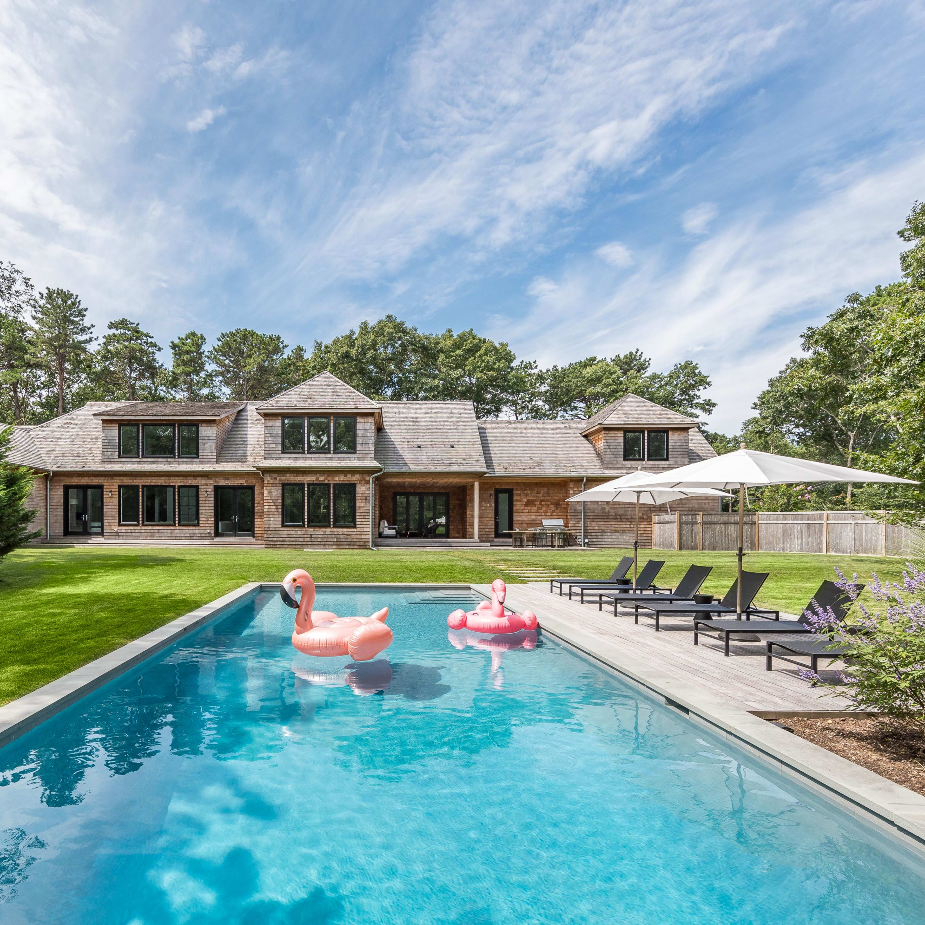 A modern house with a shingle roof is situated next to a swimming pool featuring pink flamingo floats, surrounded by a well-maintained lawn and lounging chairs.