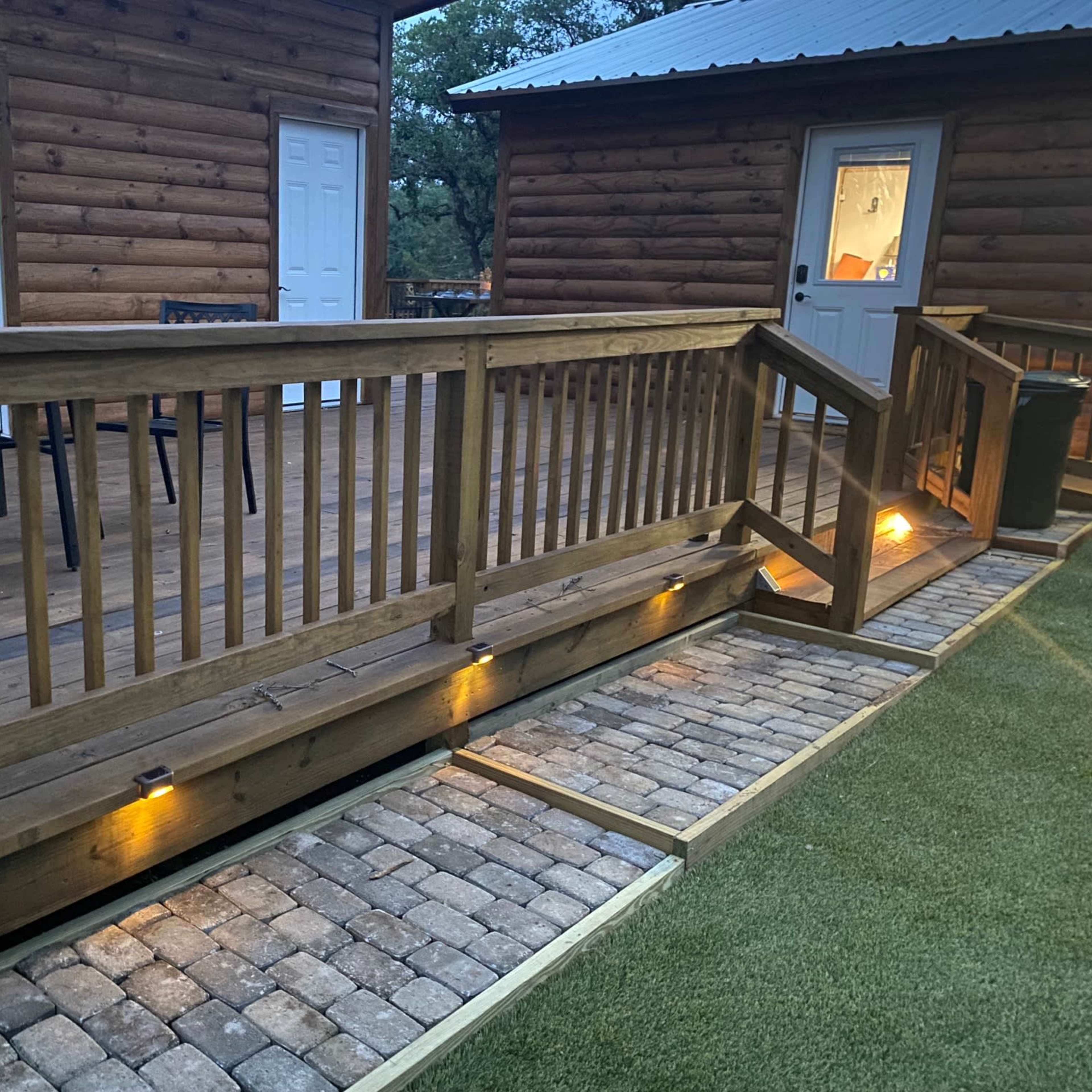 A wooden deck with a ramp leading to two cabin-like structures, illuminated by lights along a paved walkway.