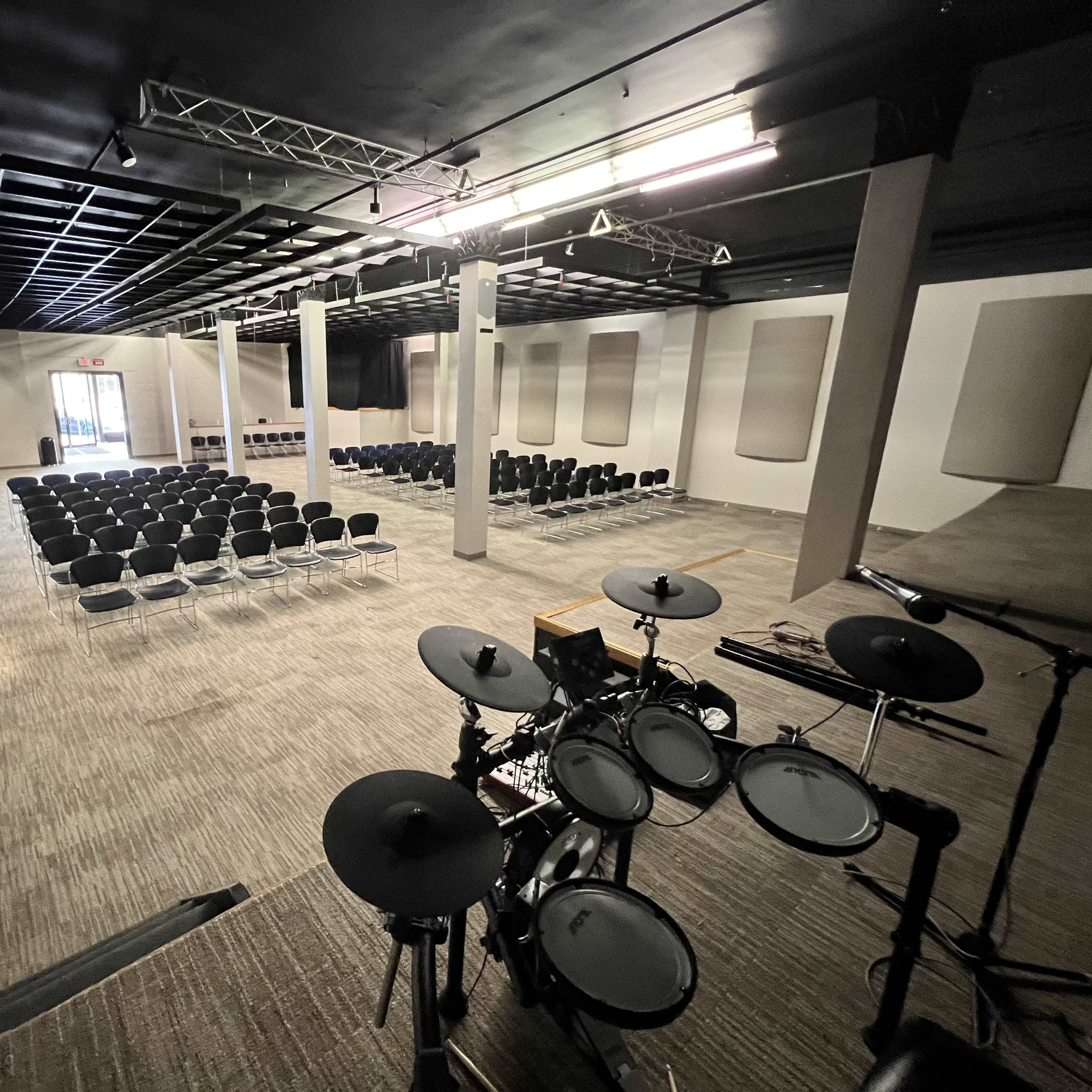 A spacious auditorium with rows of black chairs facing a stage area equipped with an electronic drum set.