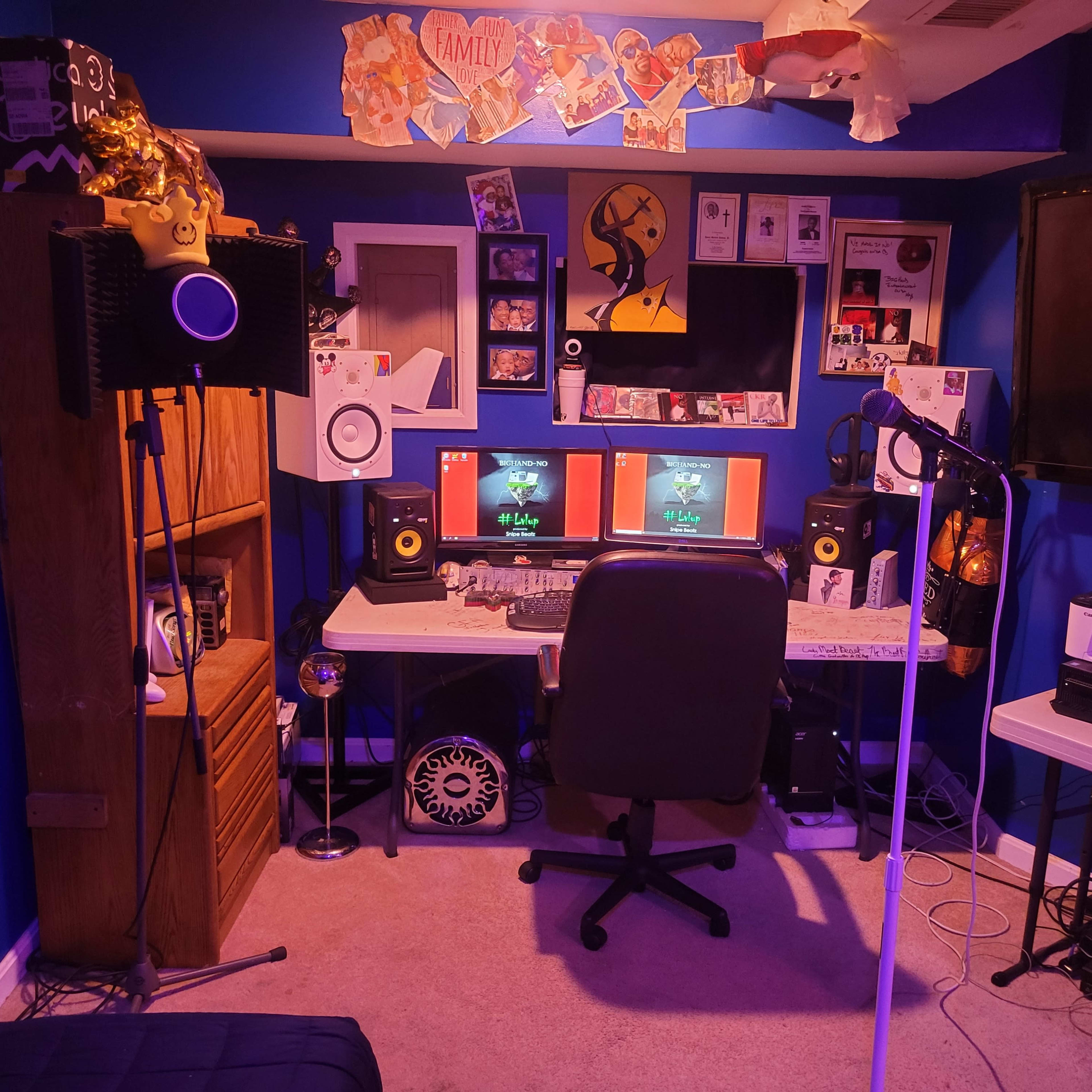 A home recording studio with two computer monitors, audio equipment, a microphone stand, and various decorations on the walls.