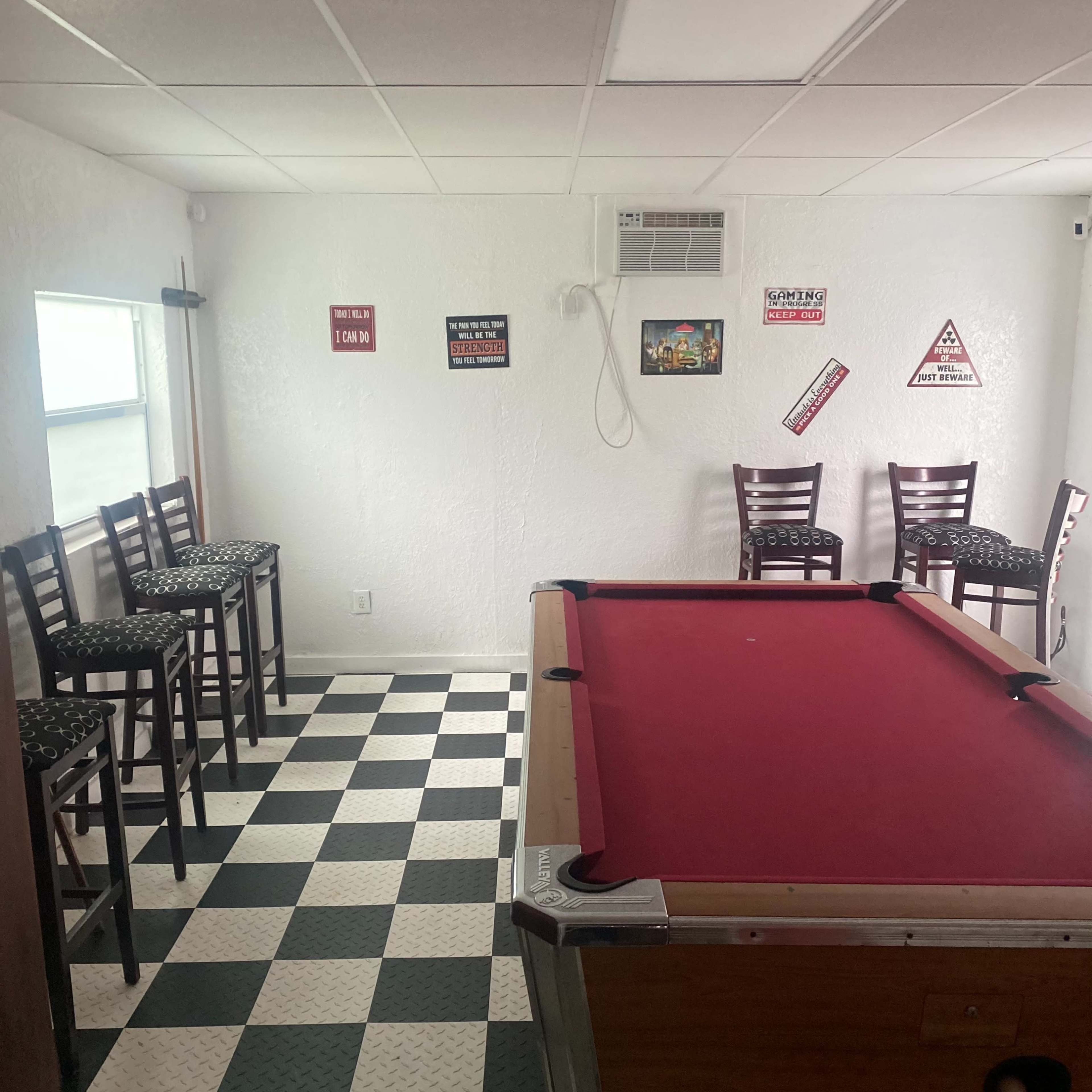 A room with a red pool table in the center, surrounded by black and white checkered flooring and several high-backed chairs against the walls.