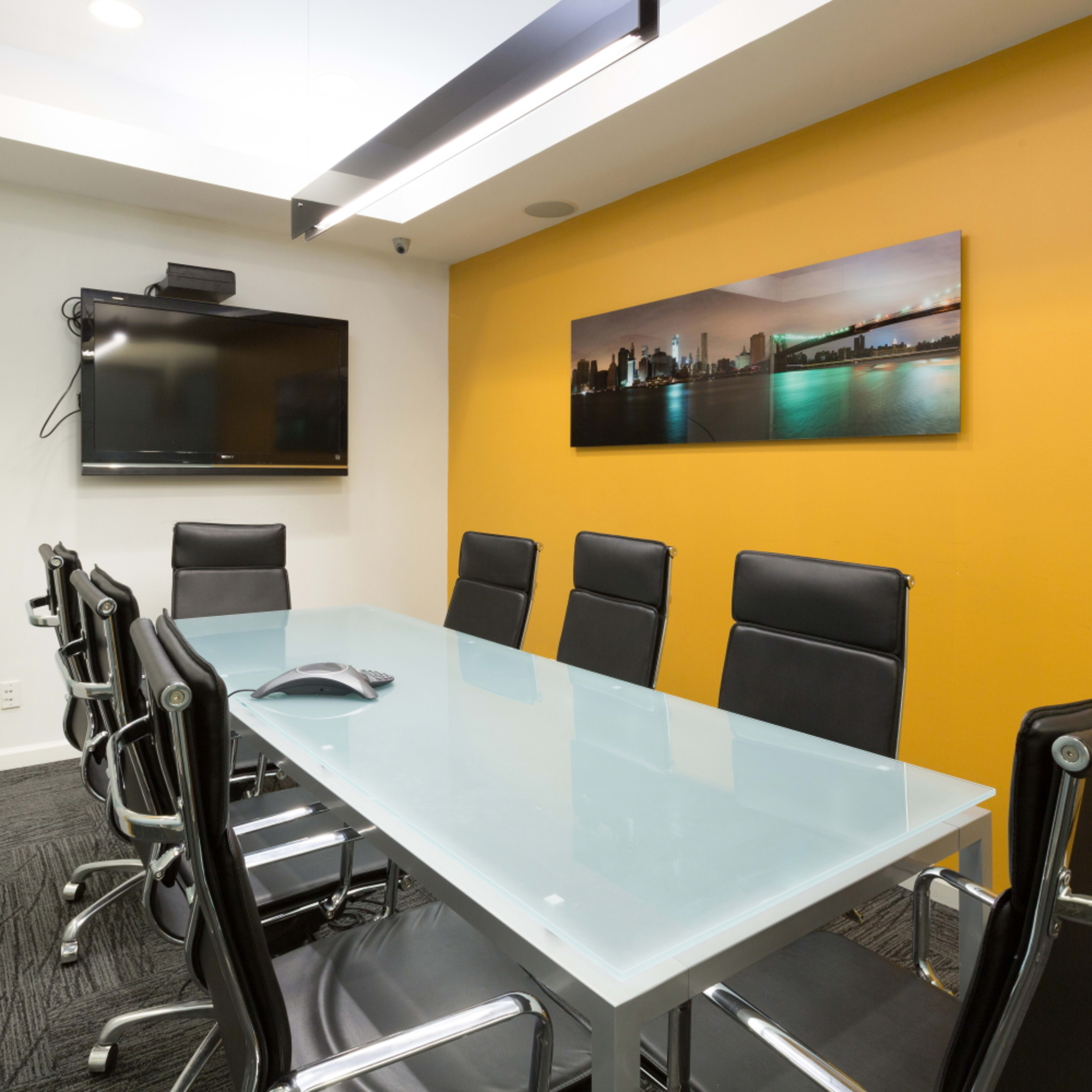 A conference room features a glass table surrounded by black chairs, with a wall-mounted TV and a vibrant orange accent wall.