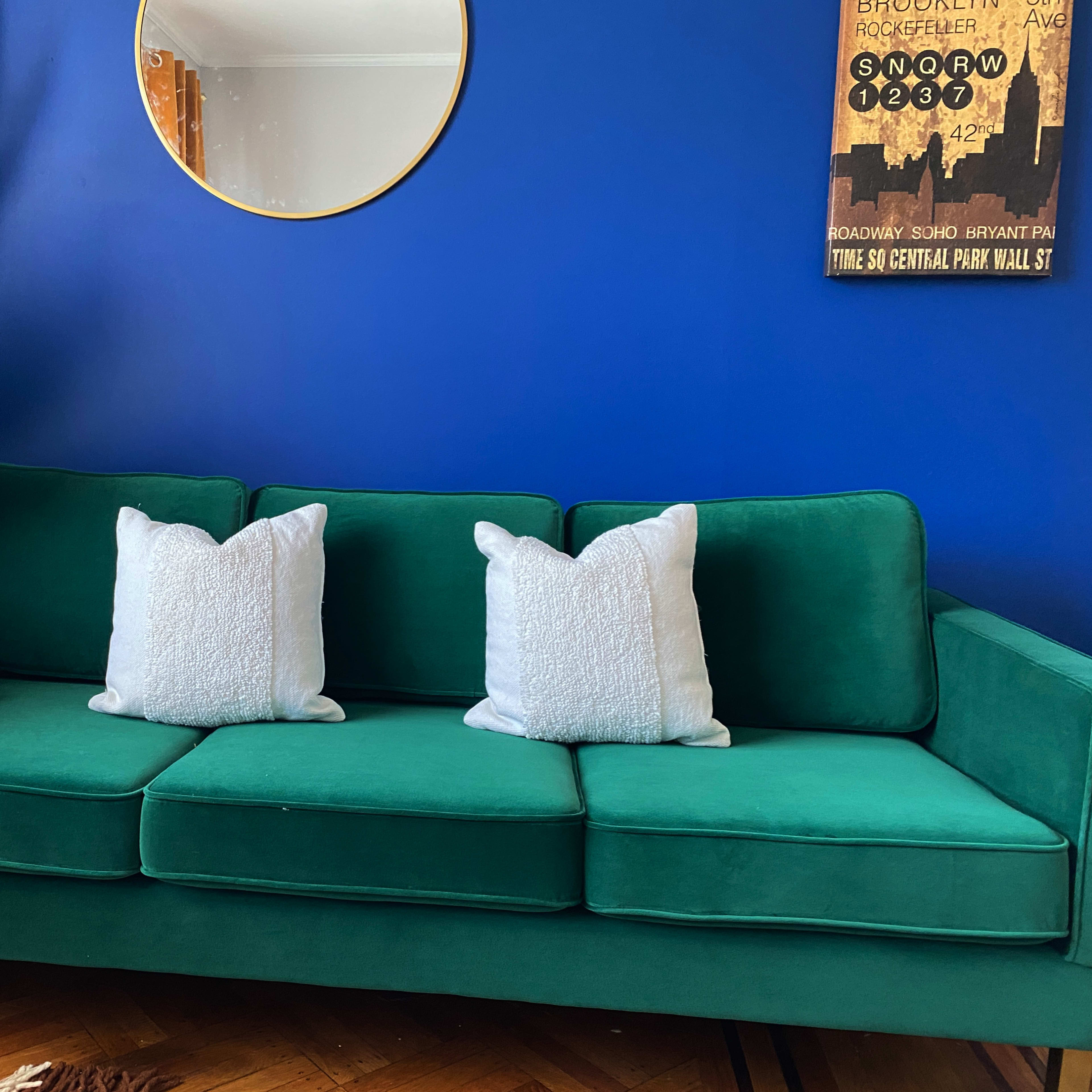 A green sofa with two white pillows, placed against a blue wall with a round mirror and a framed poster.