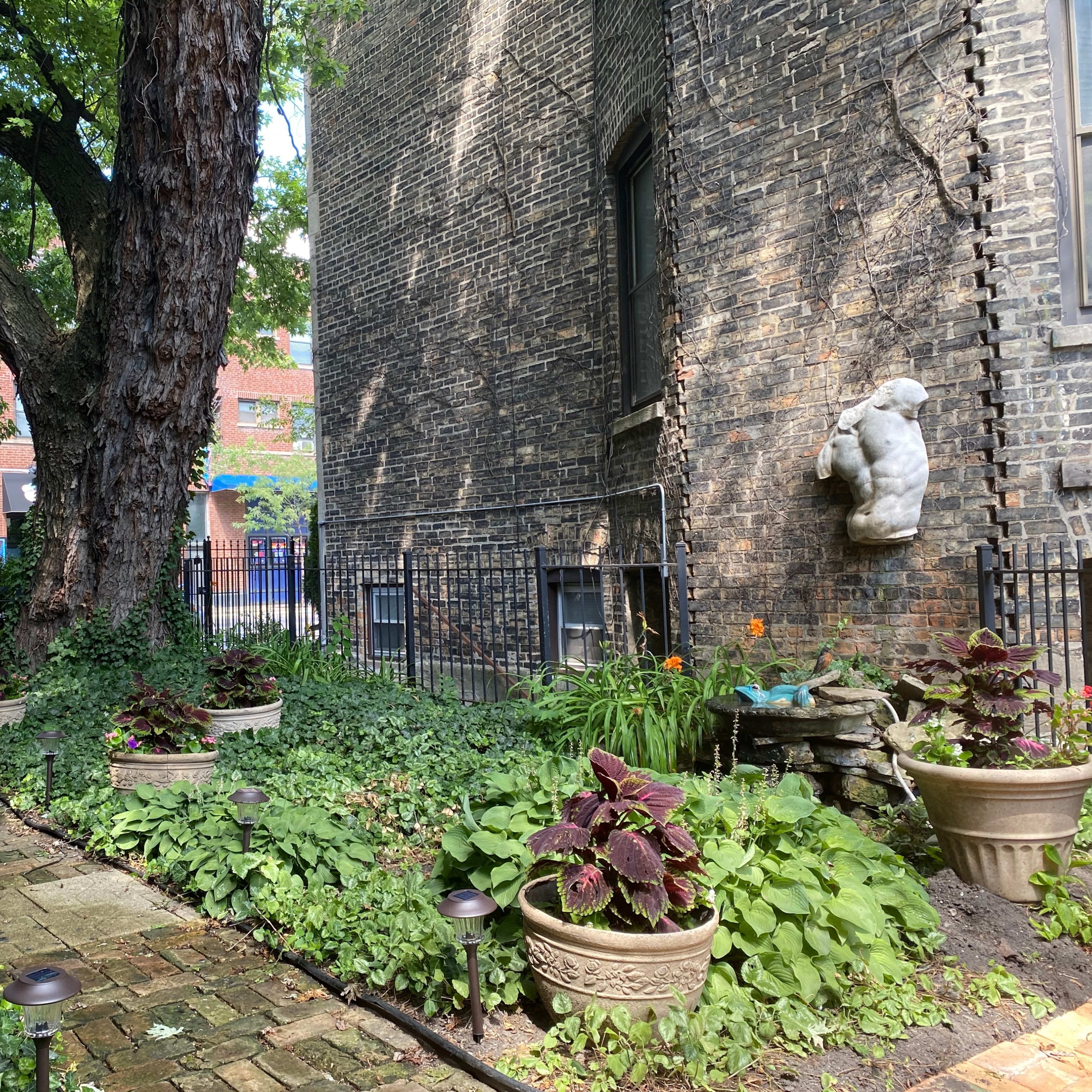 A stone sculpture of a bear is mounted on a brick wall adjacent to a lush, landscaped garden filled with potted plants and a winding stone path.