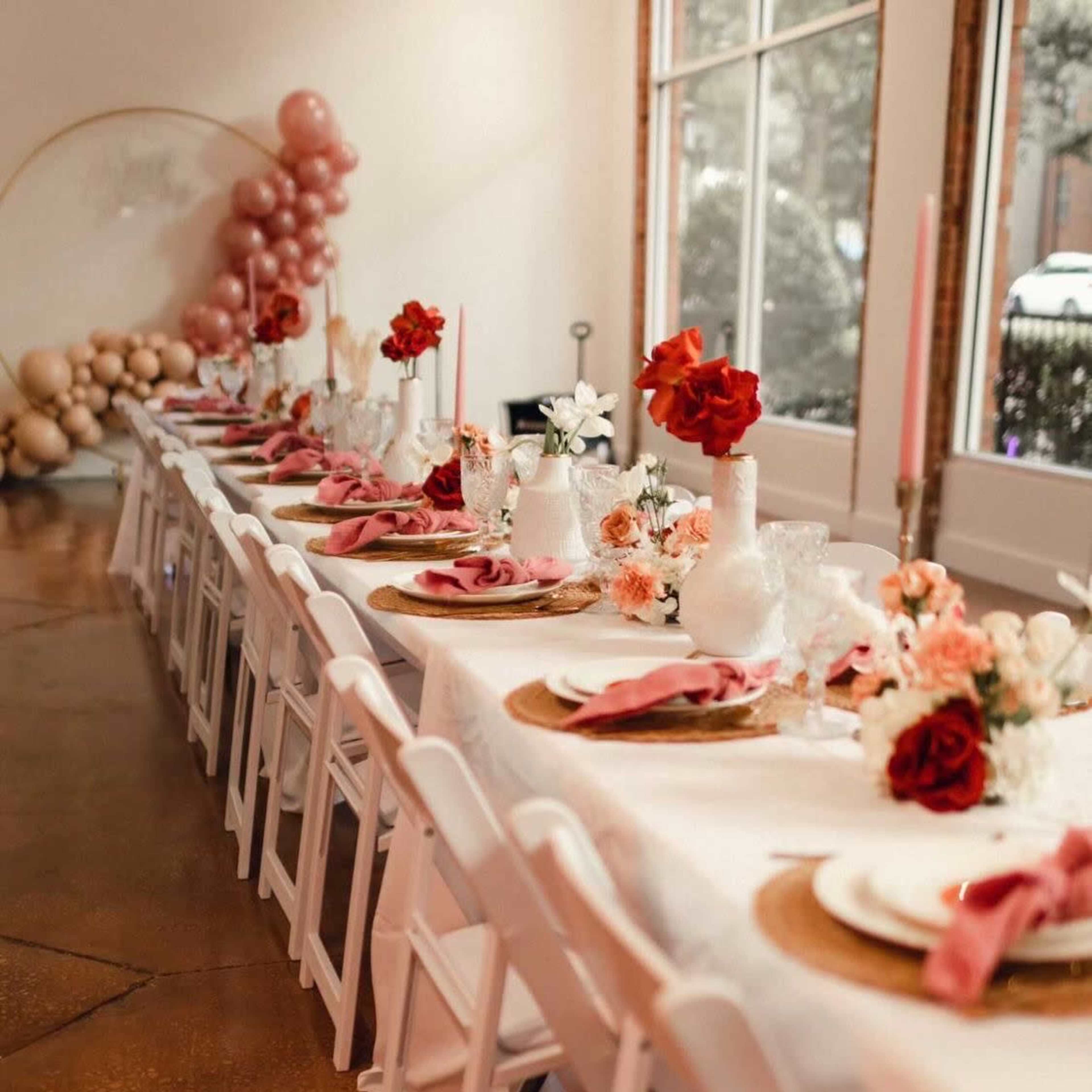 A long dining table is set with decorative plates, napkins, and floral arrangements, flanked by white chairs and a backdrop of pink balloons.